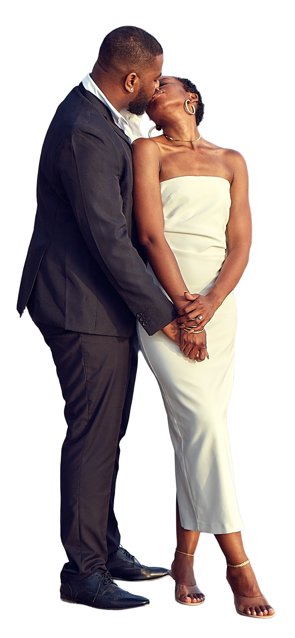 A man and woman sharing a kiss while holding hands, dressed in formal attire. The man is in a black suit and the woman is in a strapless white dress and heels.