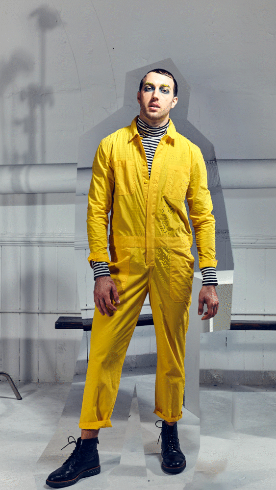Person wearing yellow coveralls and striped undershirt, standing indoors