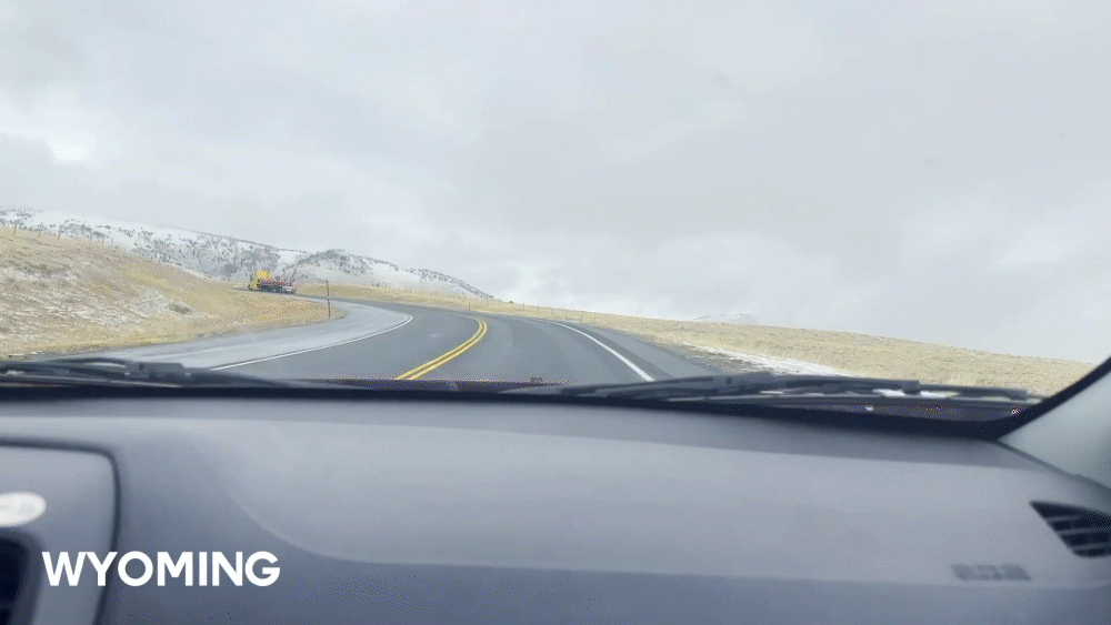 View from inside a car driving on a winding road in a snowy landscape with hills in Wyoming.