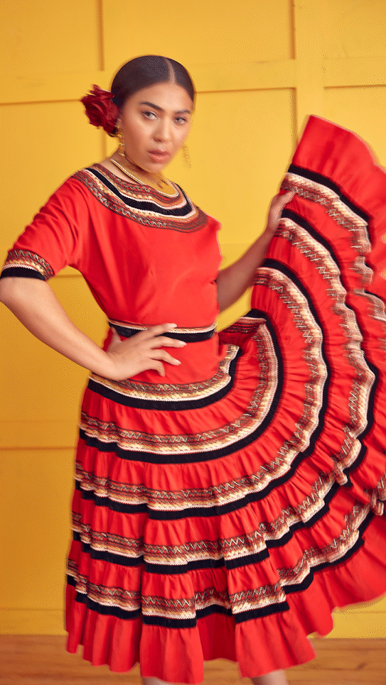A person wearing a traditional red and black dress with decorative stitching and a ruffled skirt, standing against a yellow wall. The individual has a red flower in their hair and is holding part of the dress out to the side.