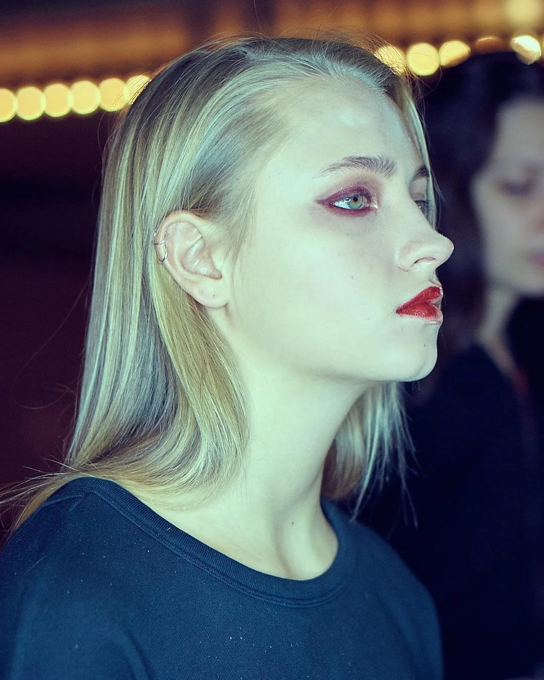 A young woman with long blonde hair, light skin, and blue eyes, wearing dark makeup with red lipstick and eyeshadow, looking to the right, in a dimly lit setting with blurred background lights.
