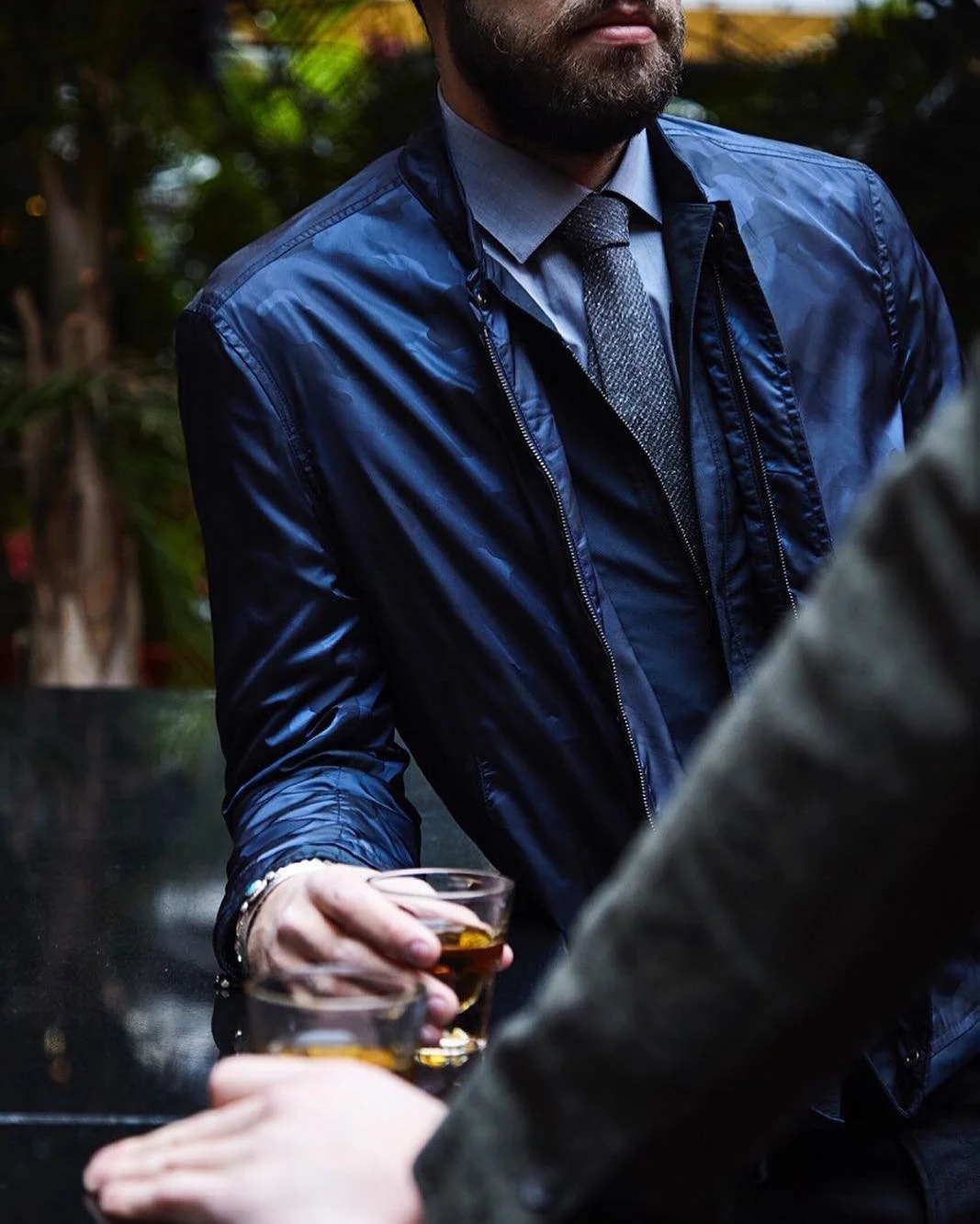 Man wearing a blue jacket and silver tie holding a glass of whiskey at an outdoor gathering, with blurred greenery in the background.