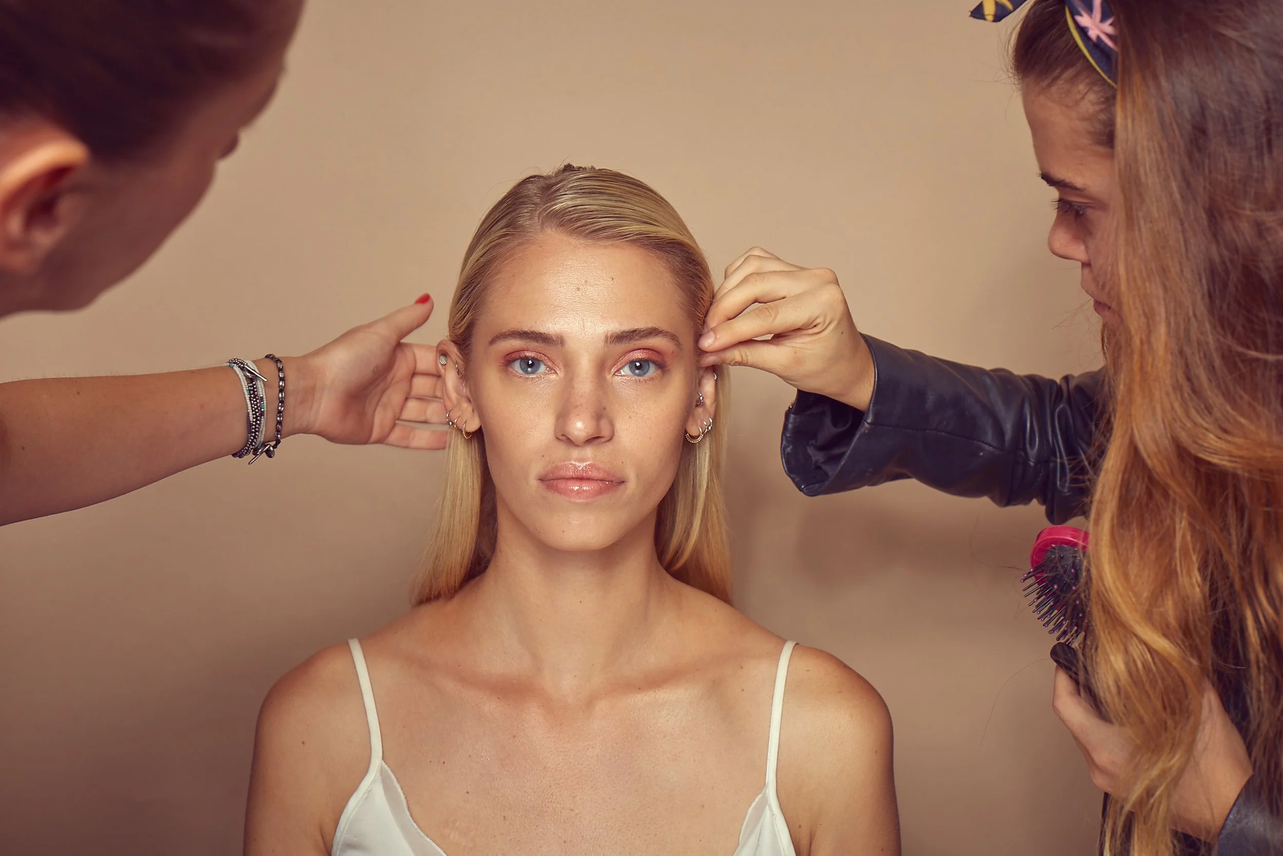 A woman with blonde hair having her makeup and hair styled by two professionals.