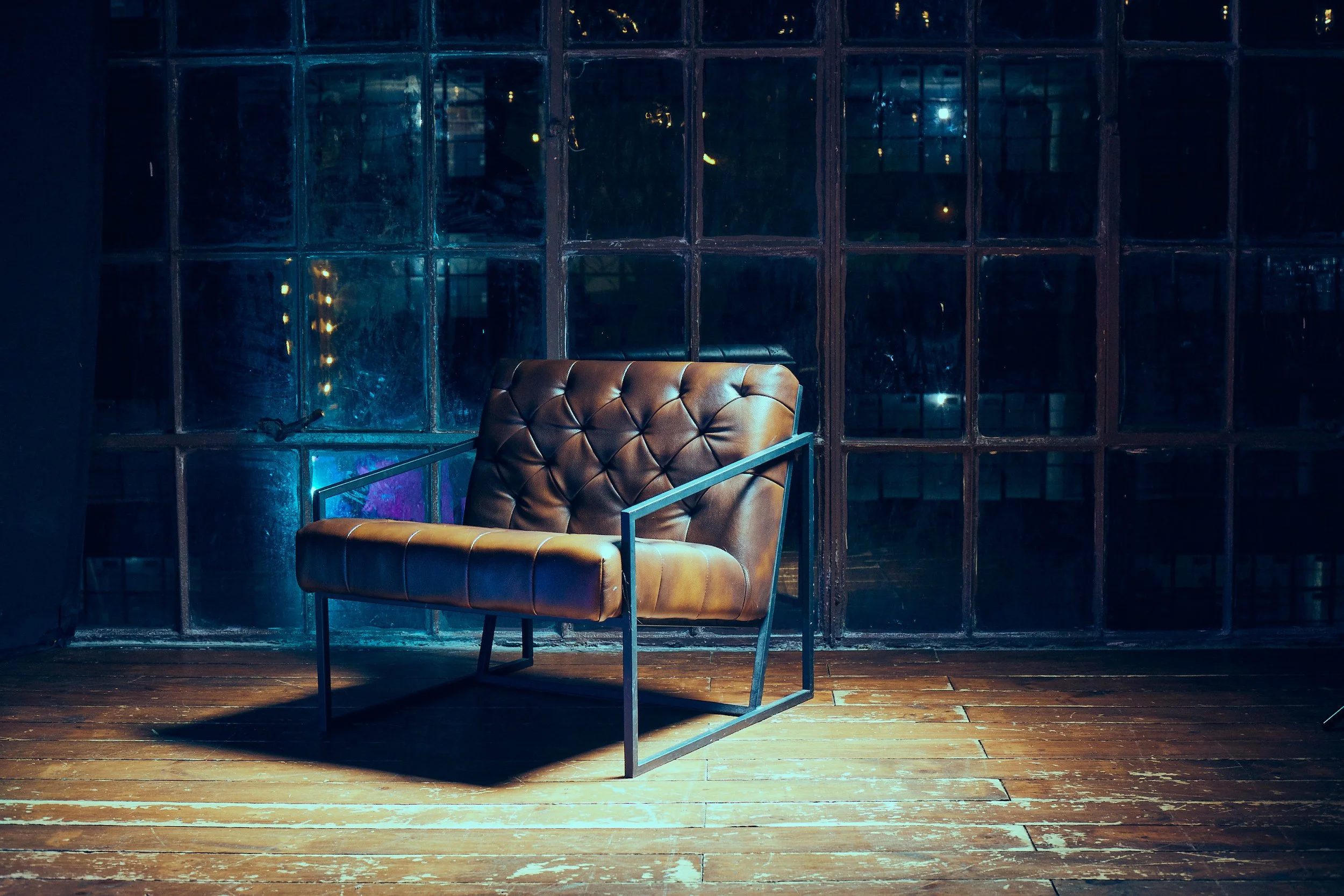 A brown leather armchair with a tufted backrest and black metal frame, placed on a wooden floor in front of a large industrial-style window with multiple panes at night.