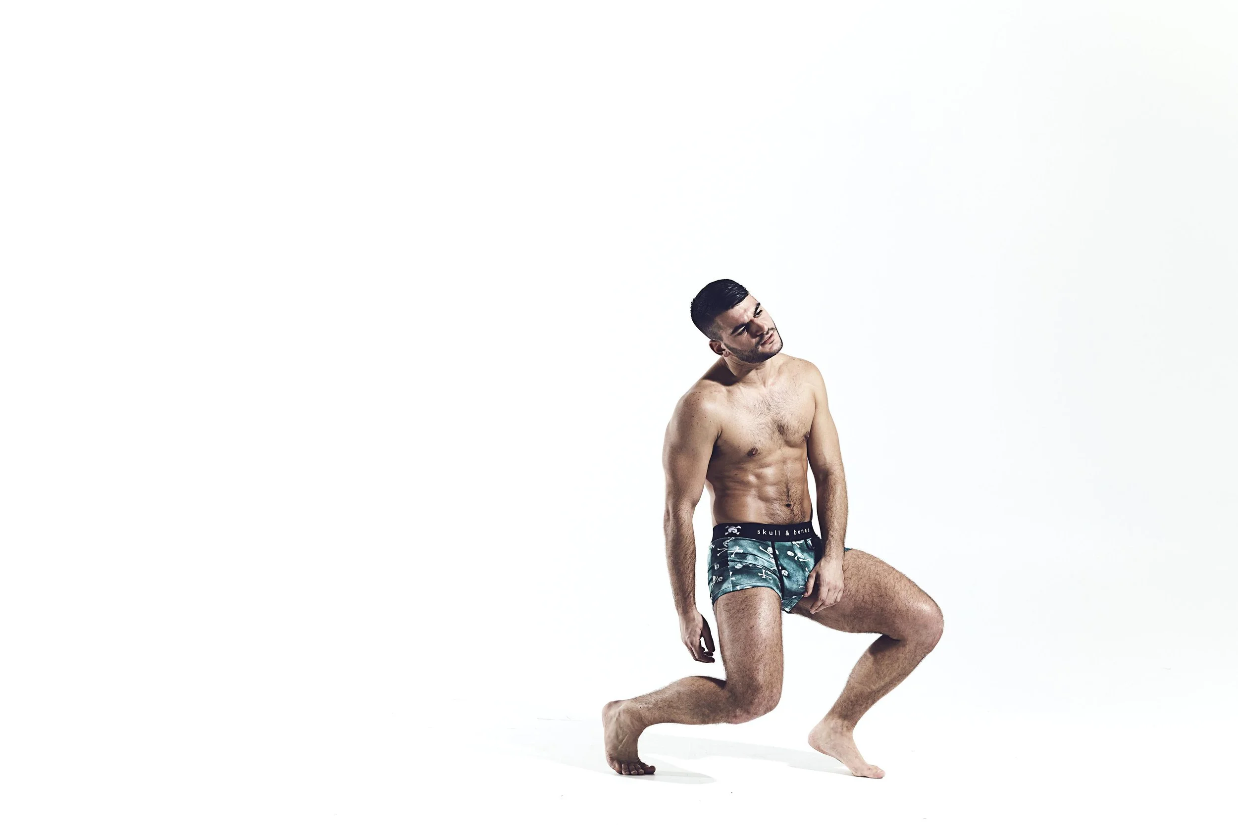 A shirtless man wearing green swim trunks in a squatting pose against a white background.