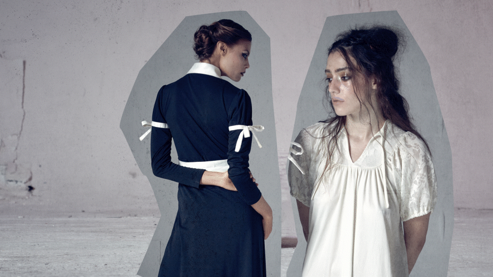 Two women standing in a minimalist setting, one in a dark dress facing away, the other in a light dress facing forward.