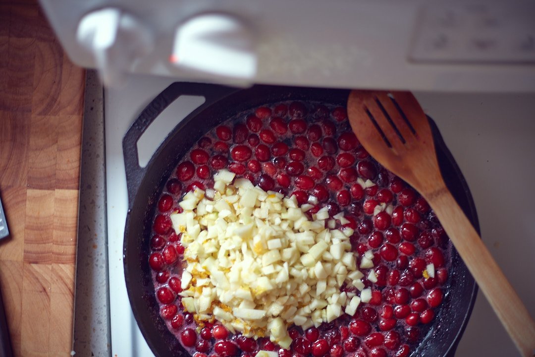 Red berries and chopped garlic cooking in a black skillet on a stove, with a wooden spatula resting inside.