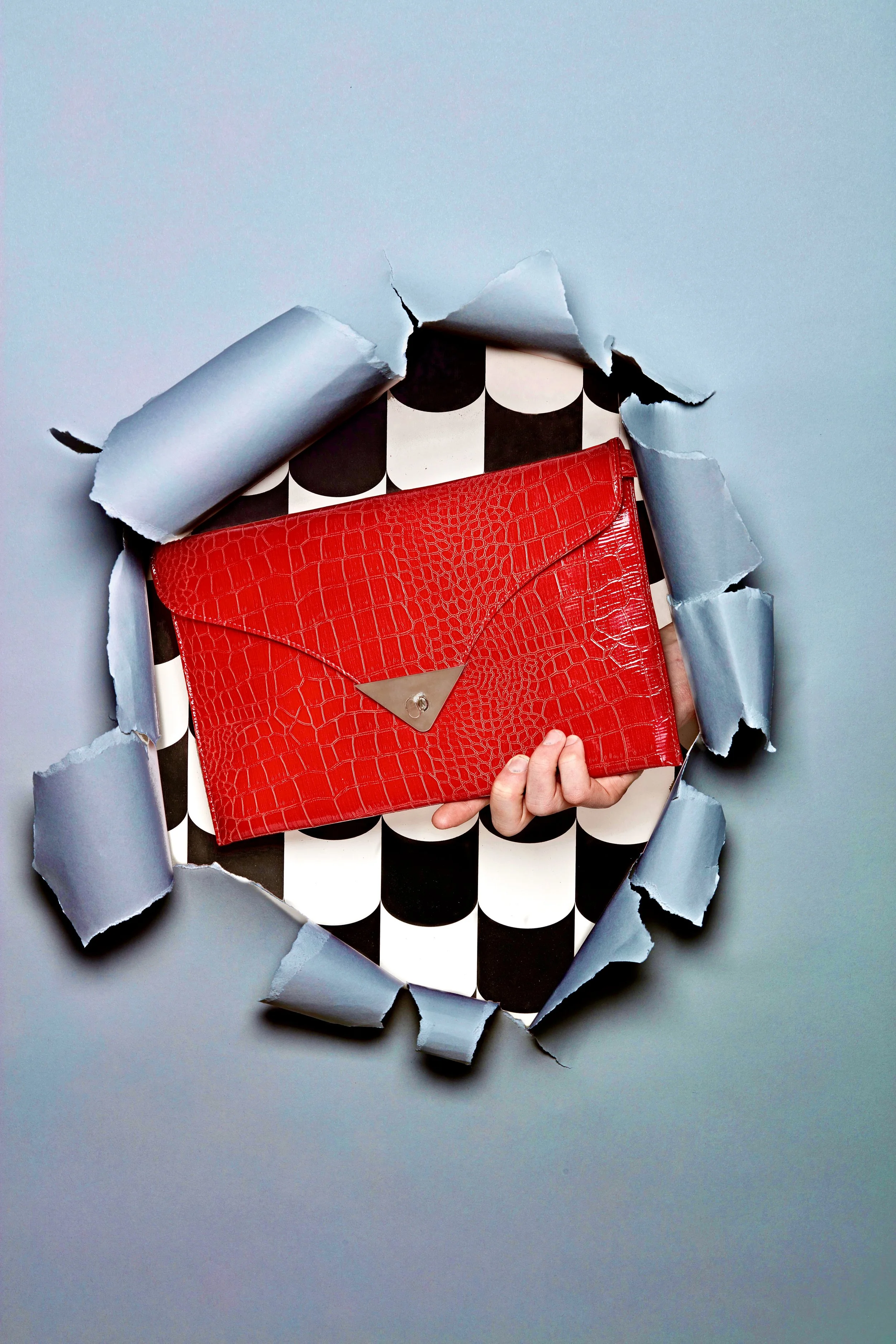 Red crocodile-embossed clutch bag with silver clasp, held through a torn hole in the wall, revealing black and white checkered interior.