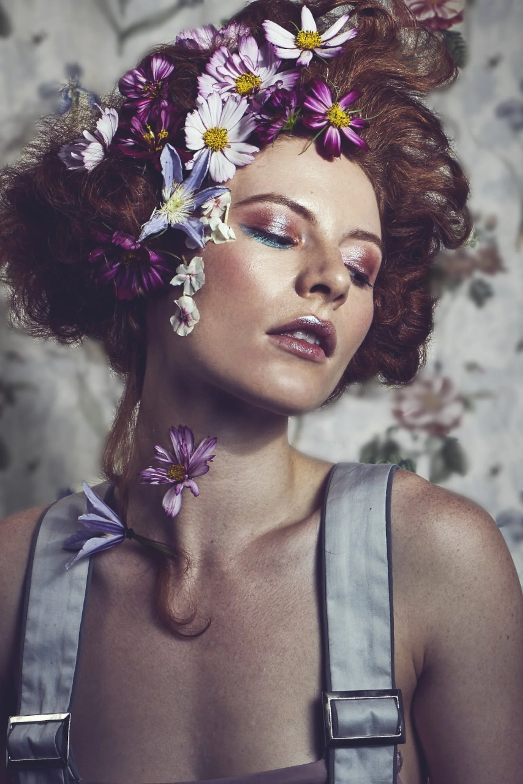 A woman with red, curly hair adorned with various pink and white flowers, with her eyes closed and wearing makeup, against a floral backdrop.