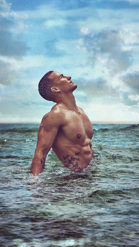 A shirtless man with tattoos on his side and chest stands in the ocean, facing upward towards a partly cloudy sky.
