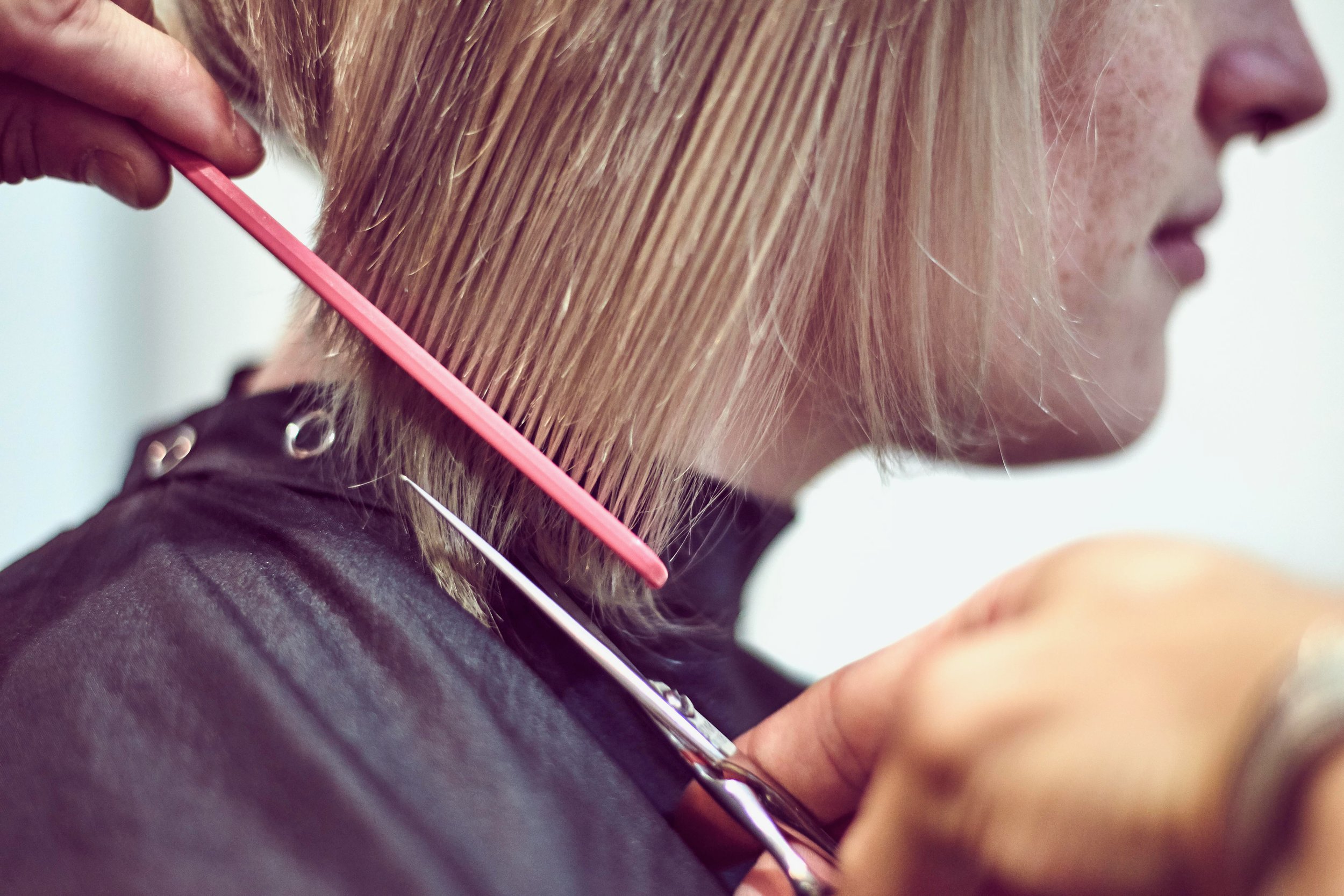 A woman getting her hair cut, with a hairstylist using scissors and a pink comb to trim her blonde hair.