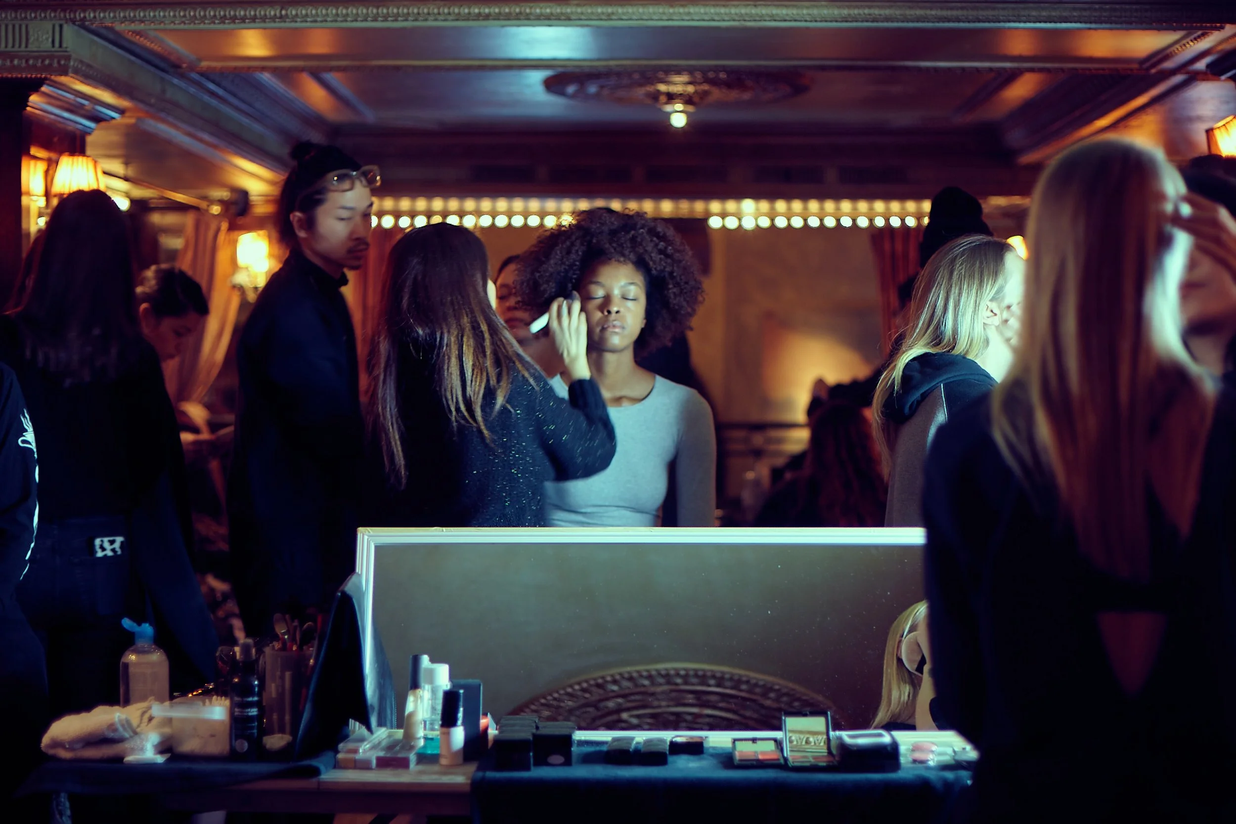 A makeup artist applies makeup to a young woman with curly hair at a beauty event, with several attendees and makeup products visible.