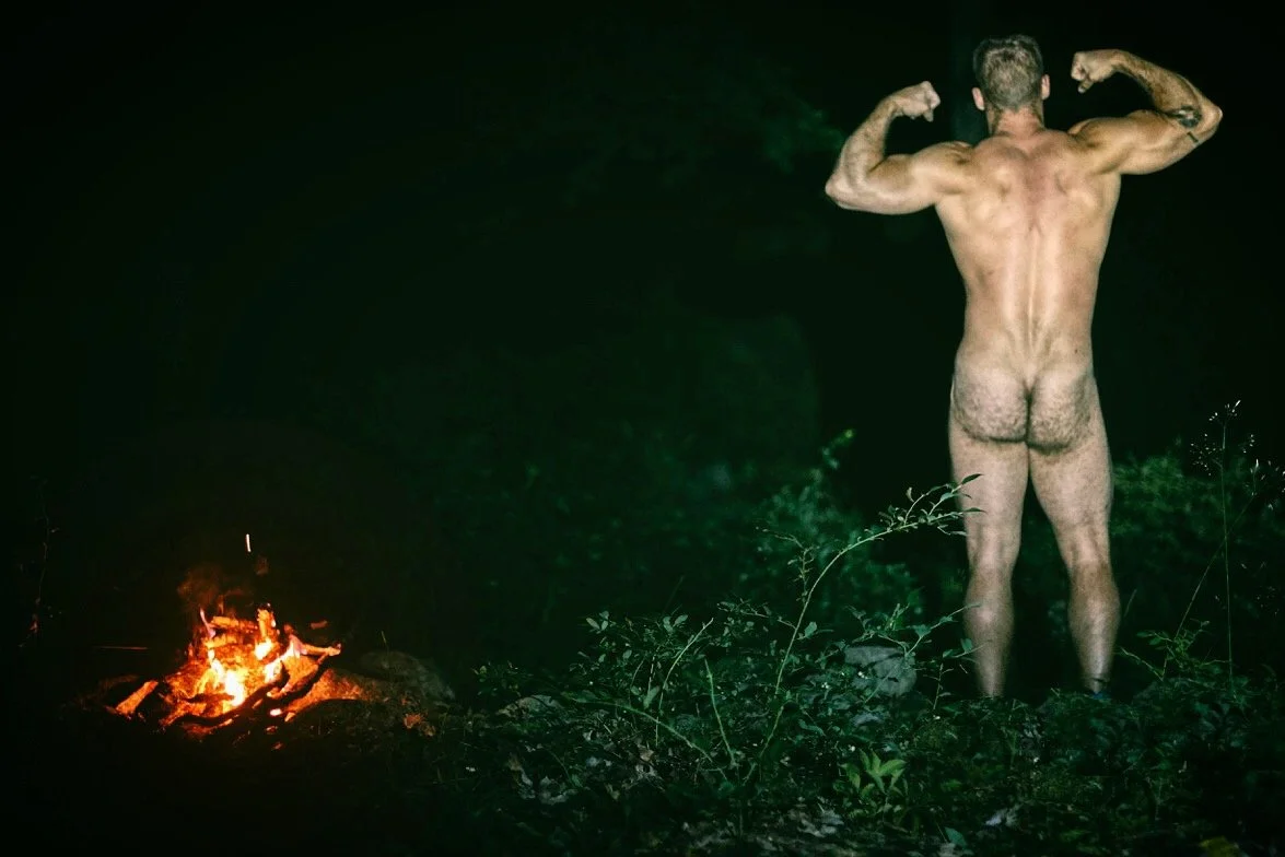 A naked man flexing muscles next to a small campfire at night.