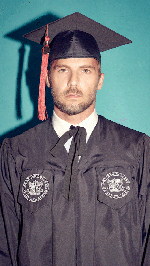 Man in graduation cap and gown standing against a blue background.