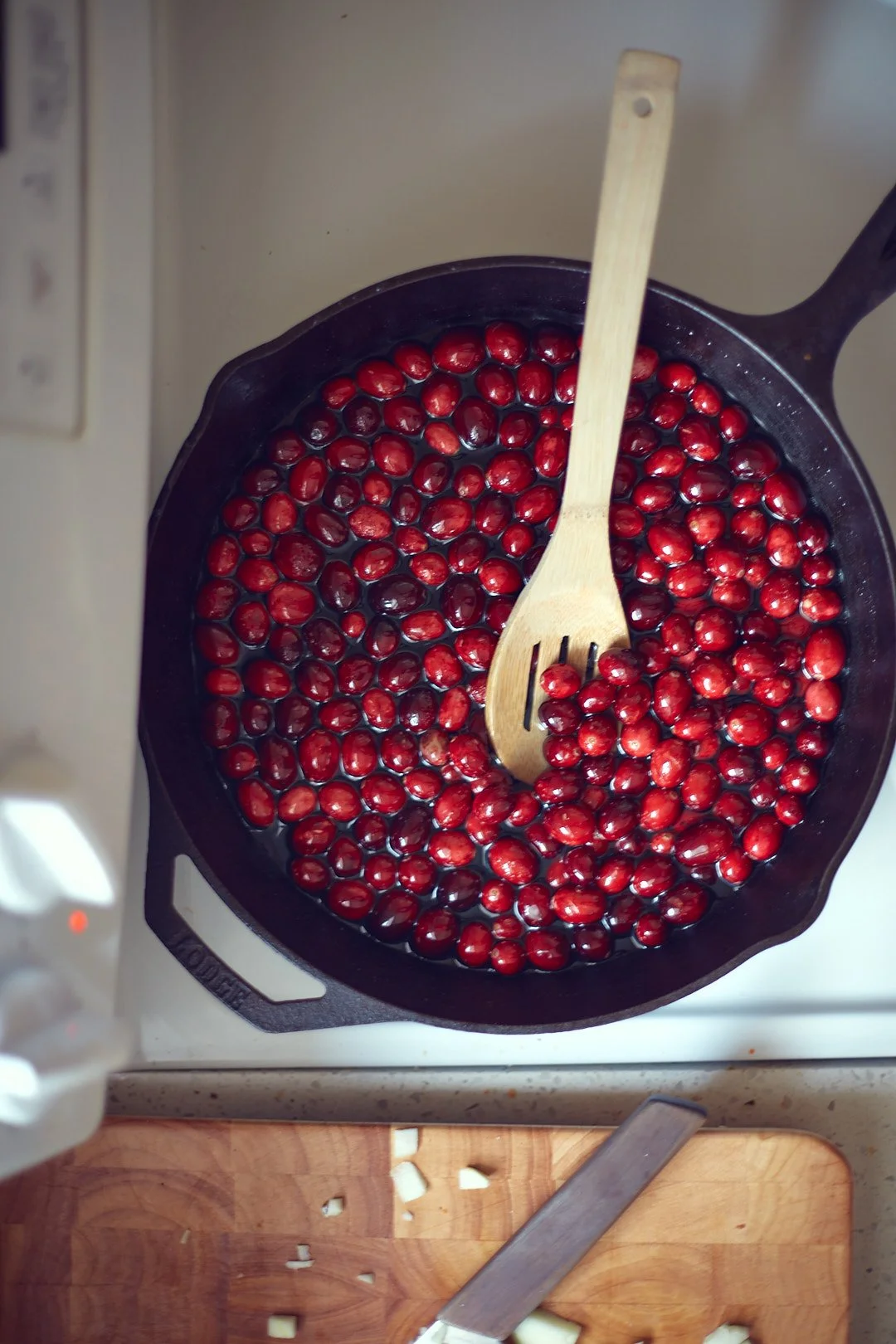 A cast iron skillet filled with fresh red cranberries, with a wooden spatula resting on top, placed on a stovetop.