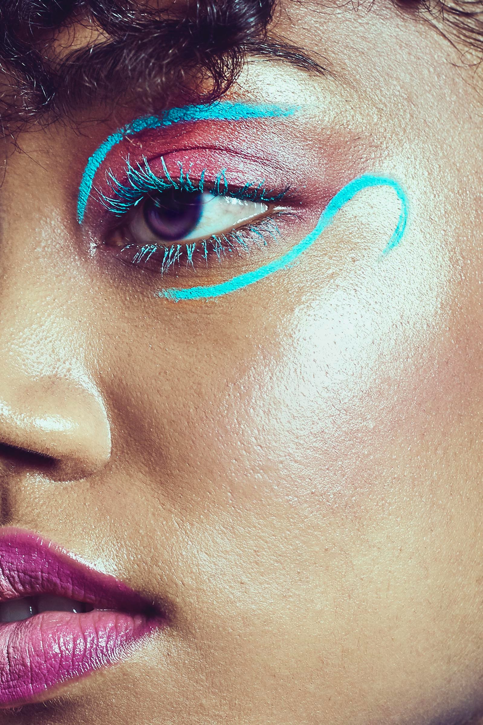 Close-up of a woman's face with vibrant makeup, featuring pink lipstick, glittery eyeshadow, and blue neon lines outlining her eyelid and cheek.