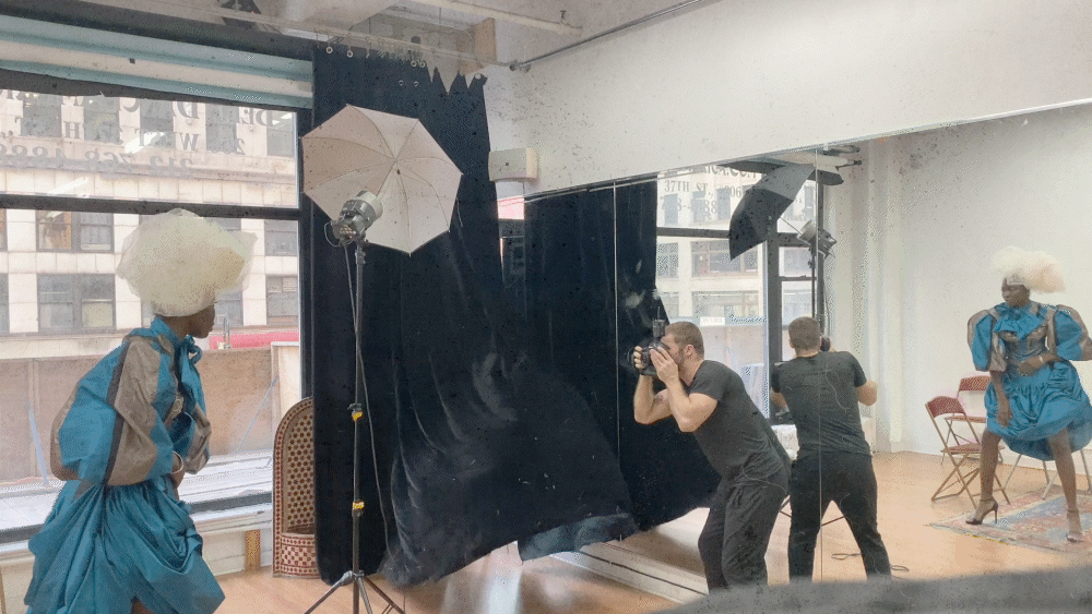 Photo shoot in a studio with photographers, models in blue outfits and large white wigs, and lighting equipment.