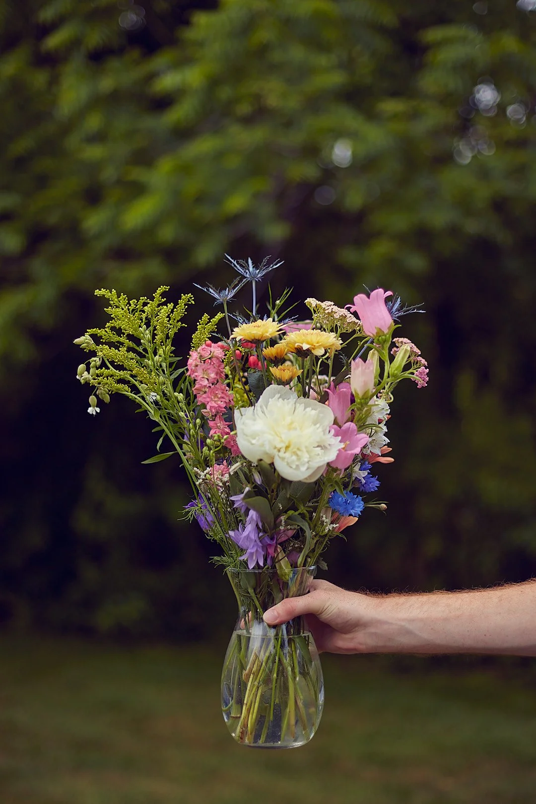 A hand holding a glass vase filled with colorful assorted flowers against a blurred outdoor background.