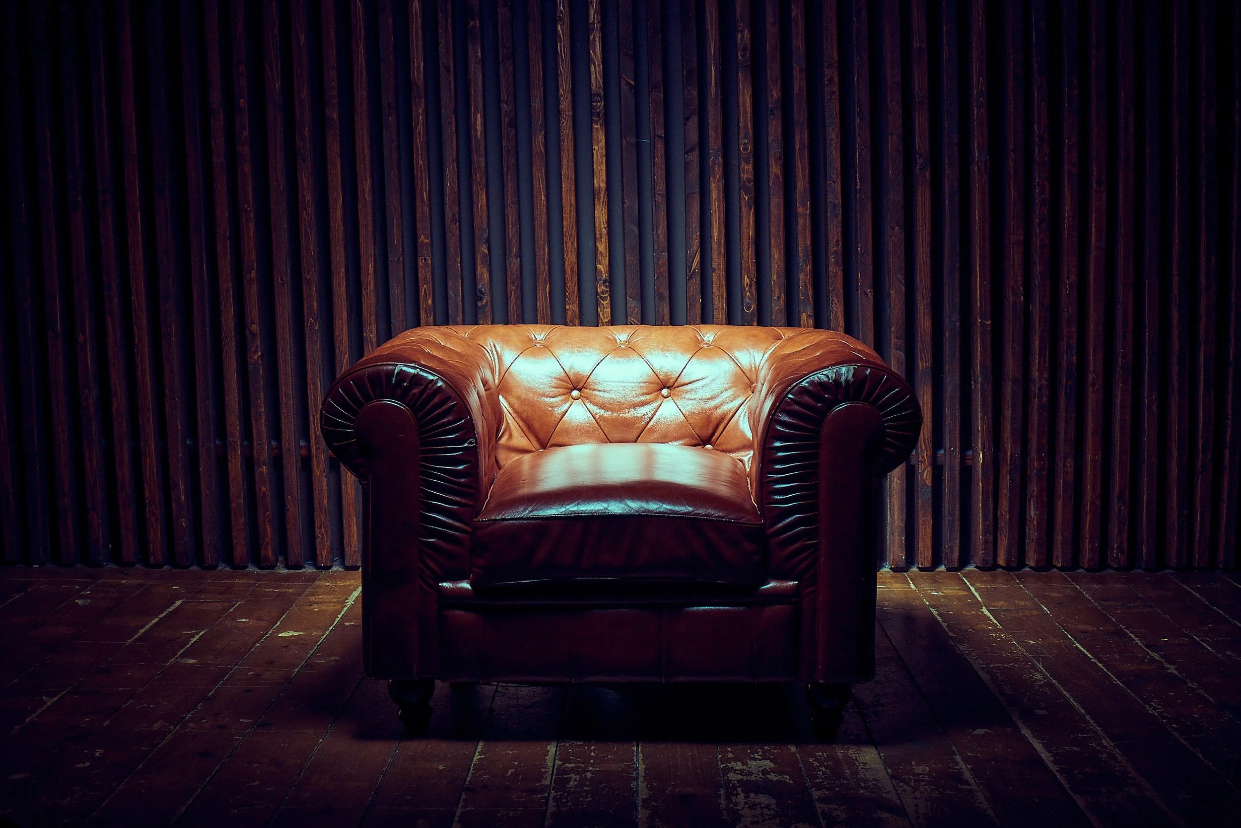 A brown leather tufted armchair with rolled arms, placed on a wooden floor against a dark wood paneled wall.