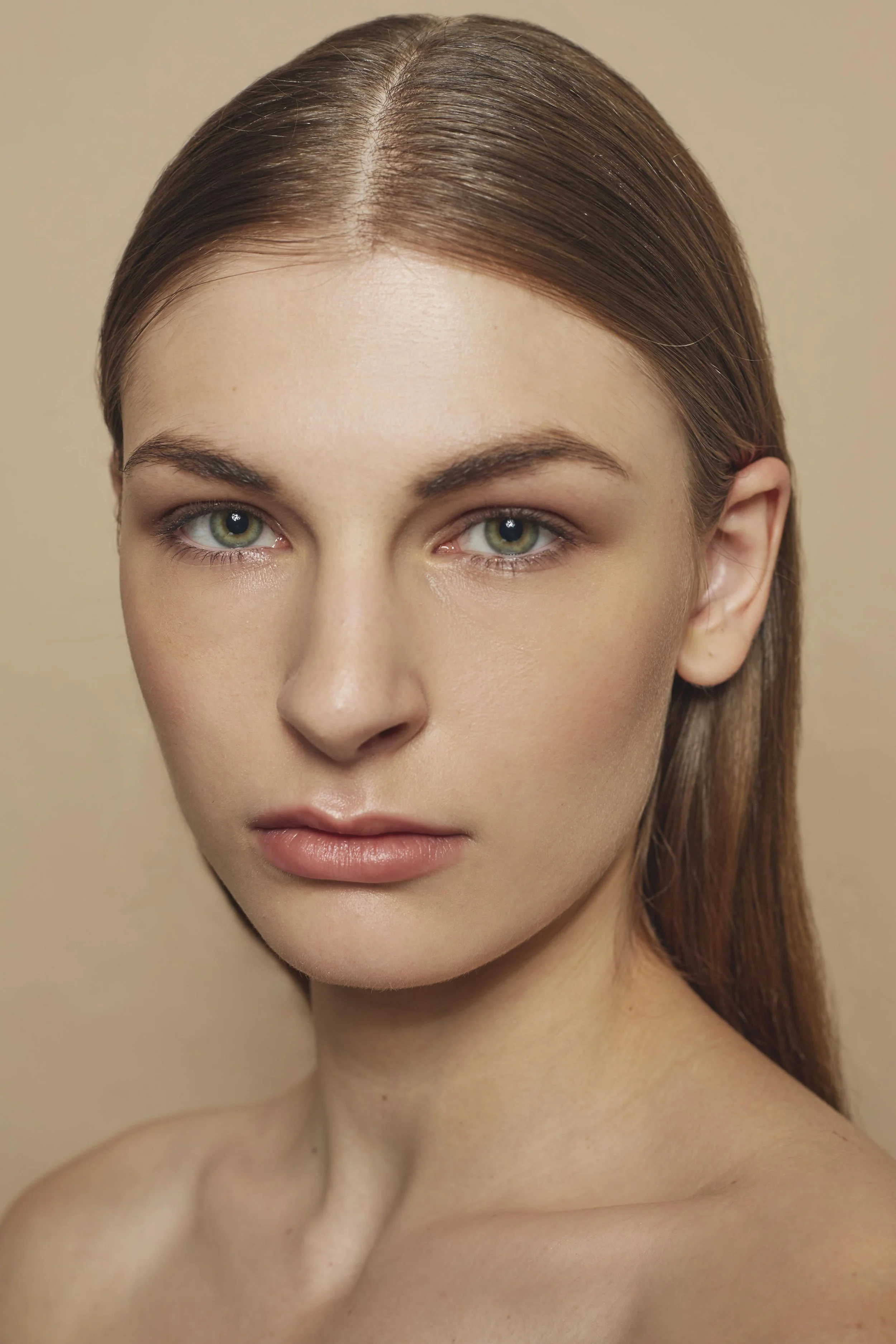 Close-up portrait of a woman with light skin, green eyes, and straight brown hair, against a beige background.