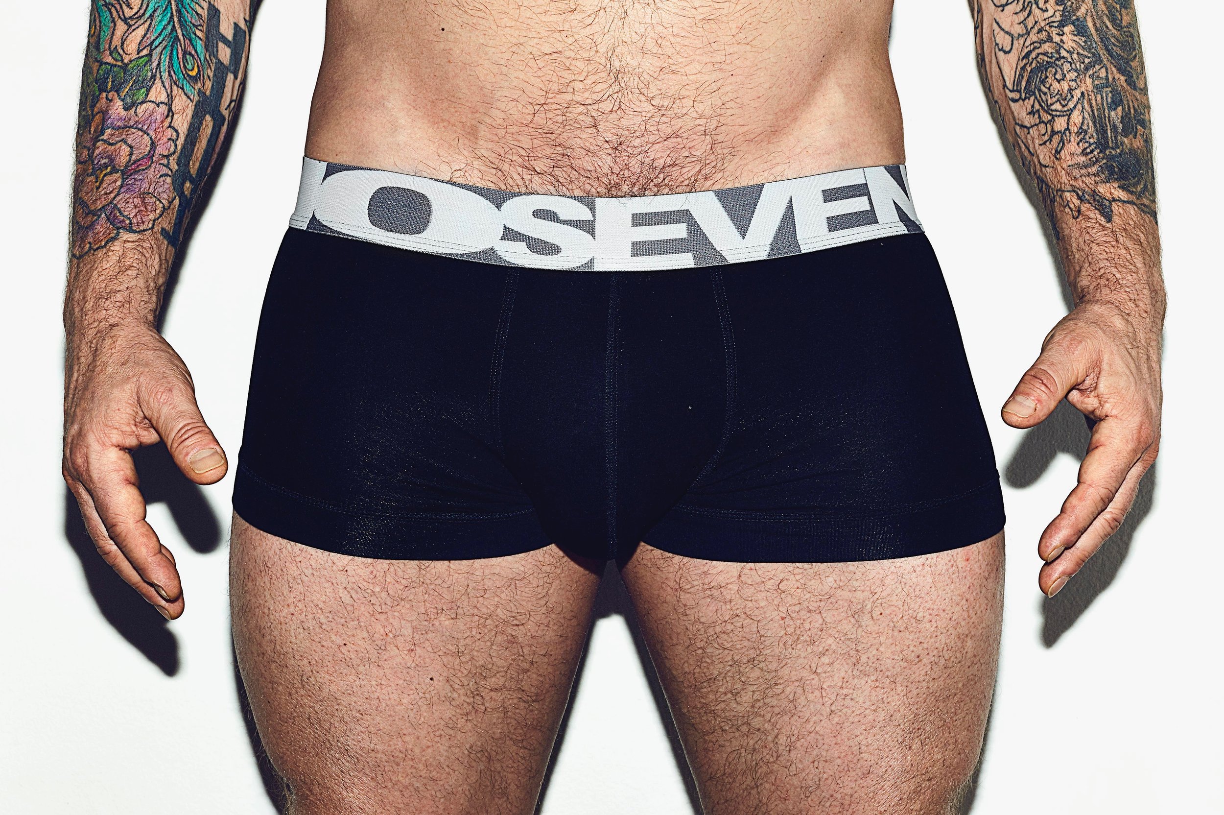 Close-up of a man's lower torso, wearing black boxer briefs with a gray waistband that has the word 'SEVEN' on it. The man has tattoos on both arms, is hairy, and has light skin.