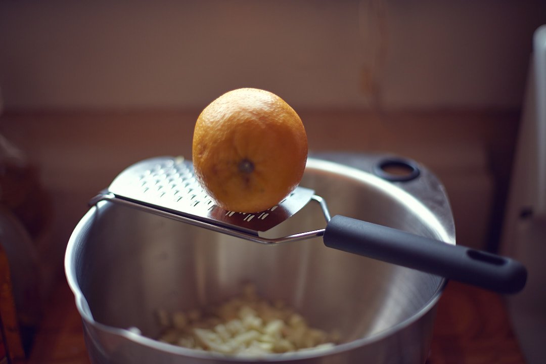A lemon is placed on a metal grater, which is resting on a metallic container or bowl. Chopped onions are visible inside the container.