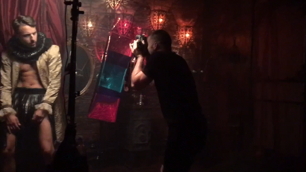 A photographer takes pictures of a male model wearing a Renaissance-style outfit in a dimly lit room with ornate decor and hanging lanterns.