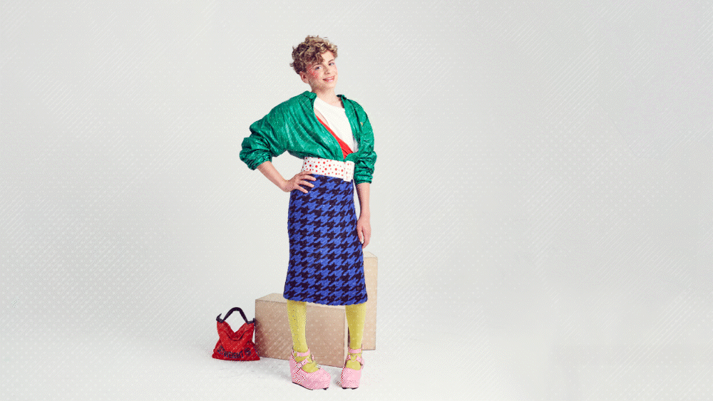 Person wearing colorful retro 80s outfit with green jacket, blue skirt, yellow tights, and pink heels, standing next to a red handbag on a neutral background.