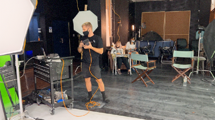 A photography studio setup with a masked photographer holding a camera, surrounded by lighting equipment and chairs. Behind, two people sit, observing the session. Various cords and accessories are scattered on the floor.