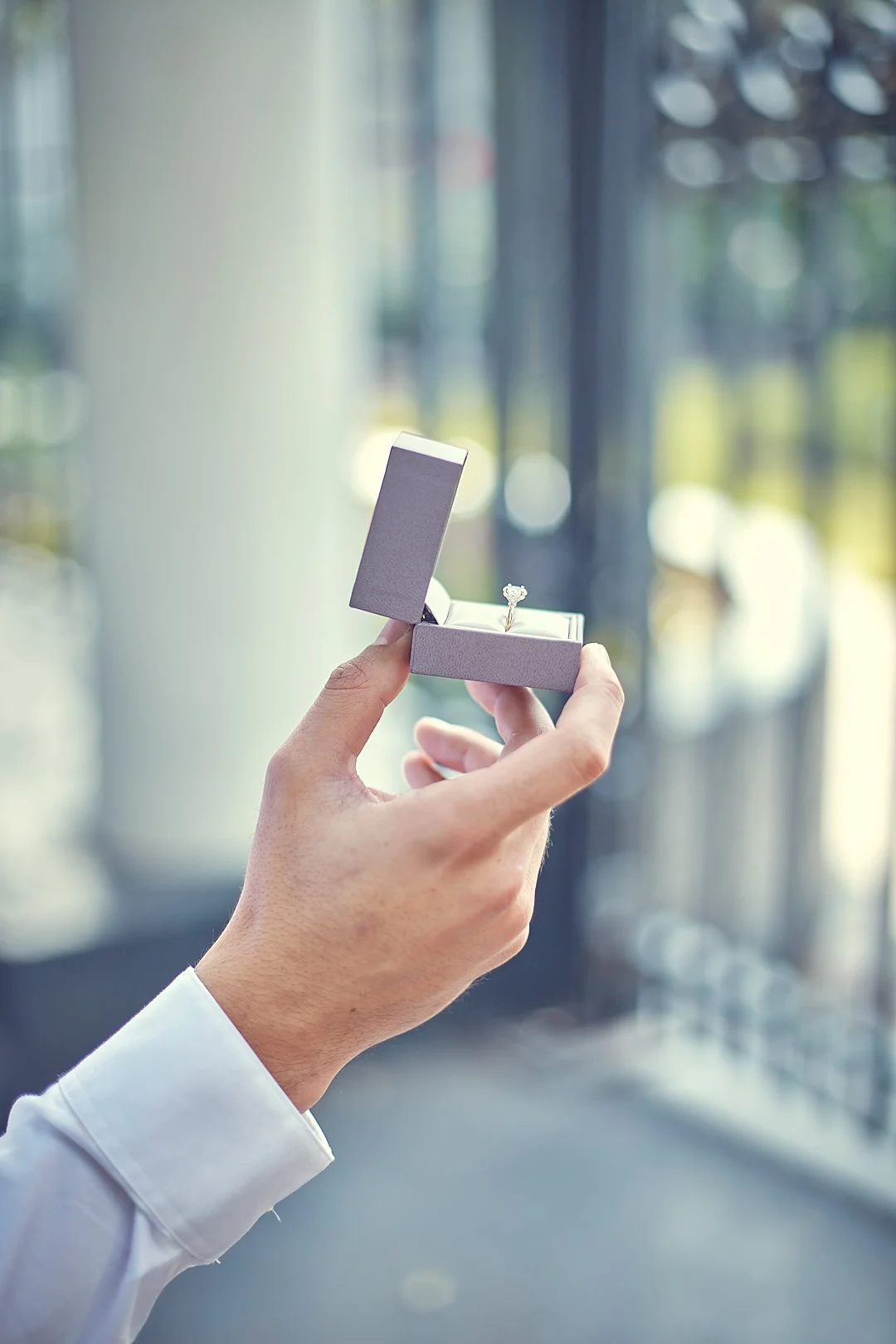 Hand holding an open jewelry box with a diamond ring inside, in front of large windows with blurred outdoor scenery.