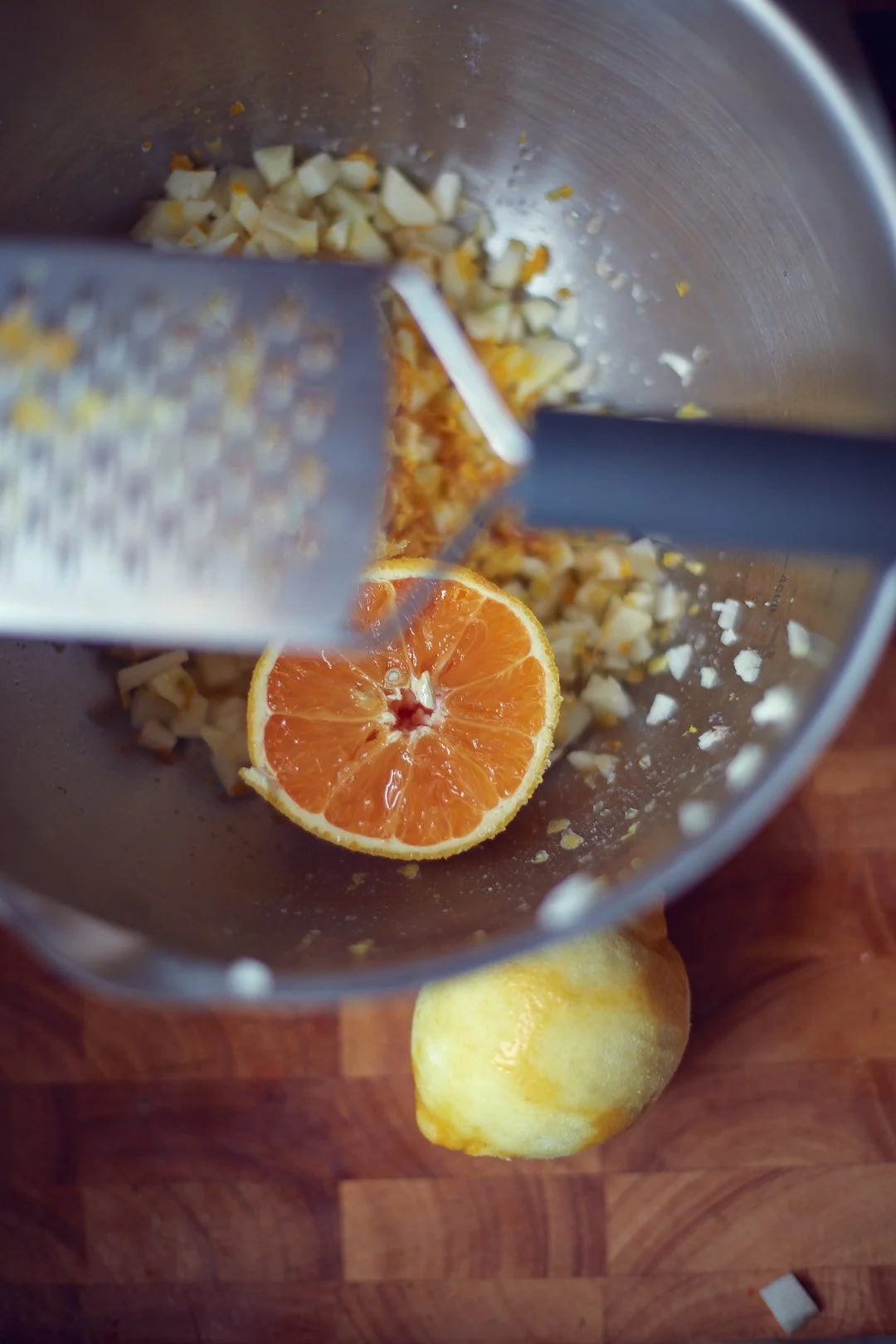 A halved orange, a lemon, chopped garlic and ginger in a blender or food processor, with a grater and wooden countertop nearby.