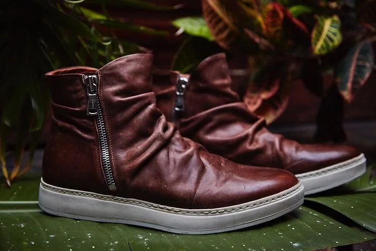 Brown leather ankle boots with zipper on the side, placed on a green leafy surface with foliage in the background.
