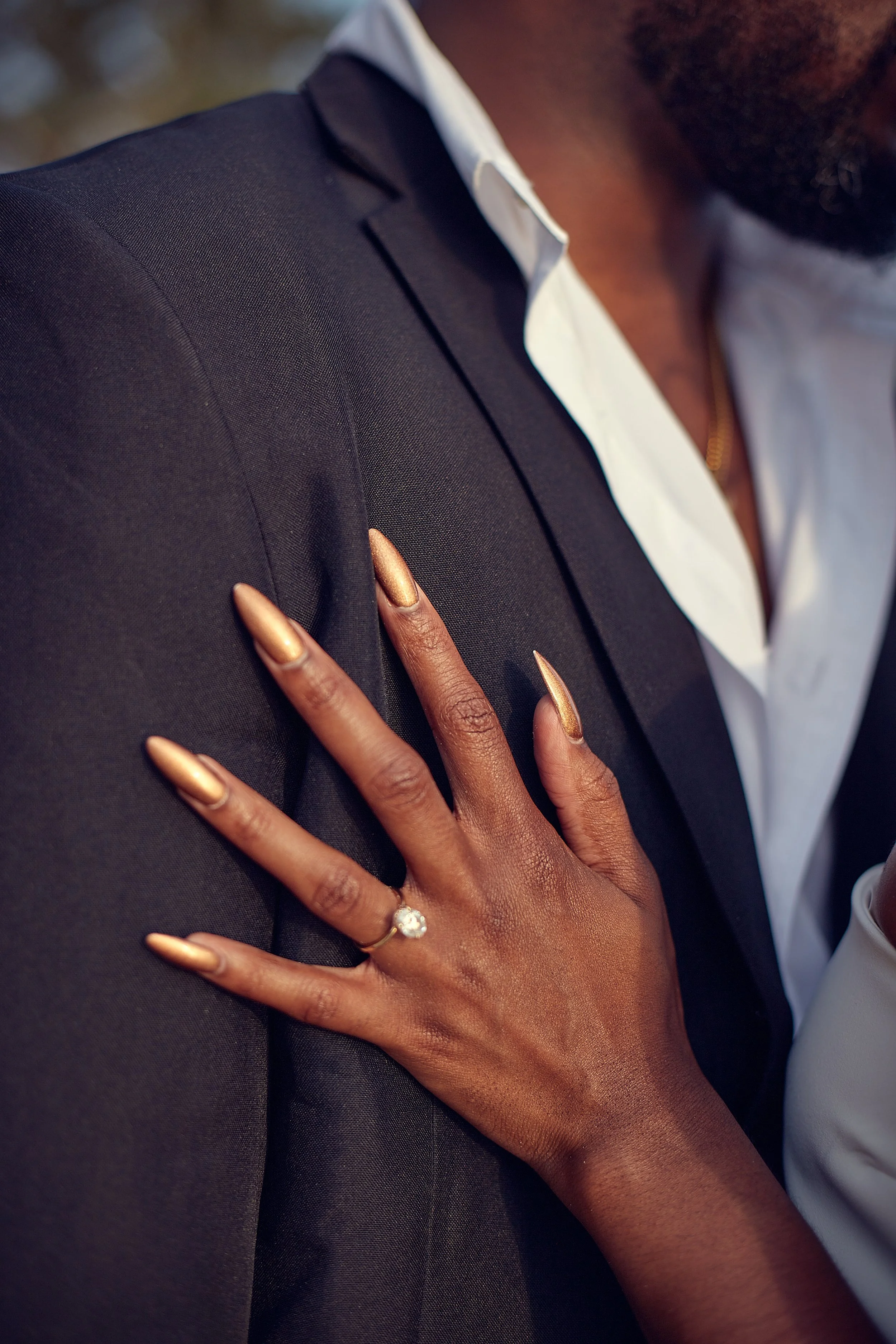 Close-up of a woman with dark skin wearing a diamond ring on her finger, with her hand resting on a man's shoulder dressed in a dark suit and white shirt.