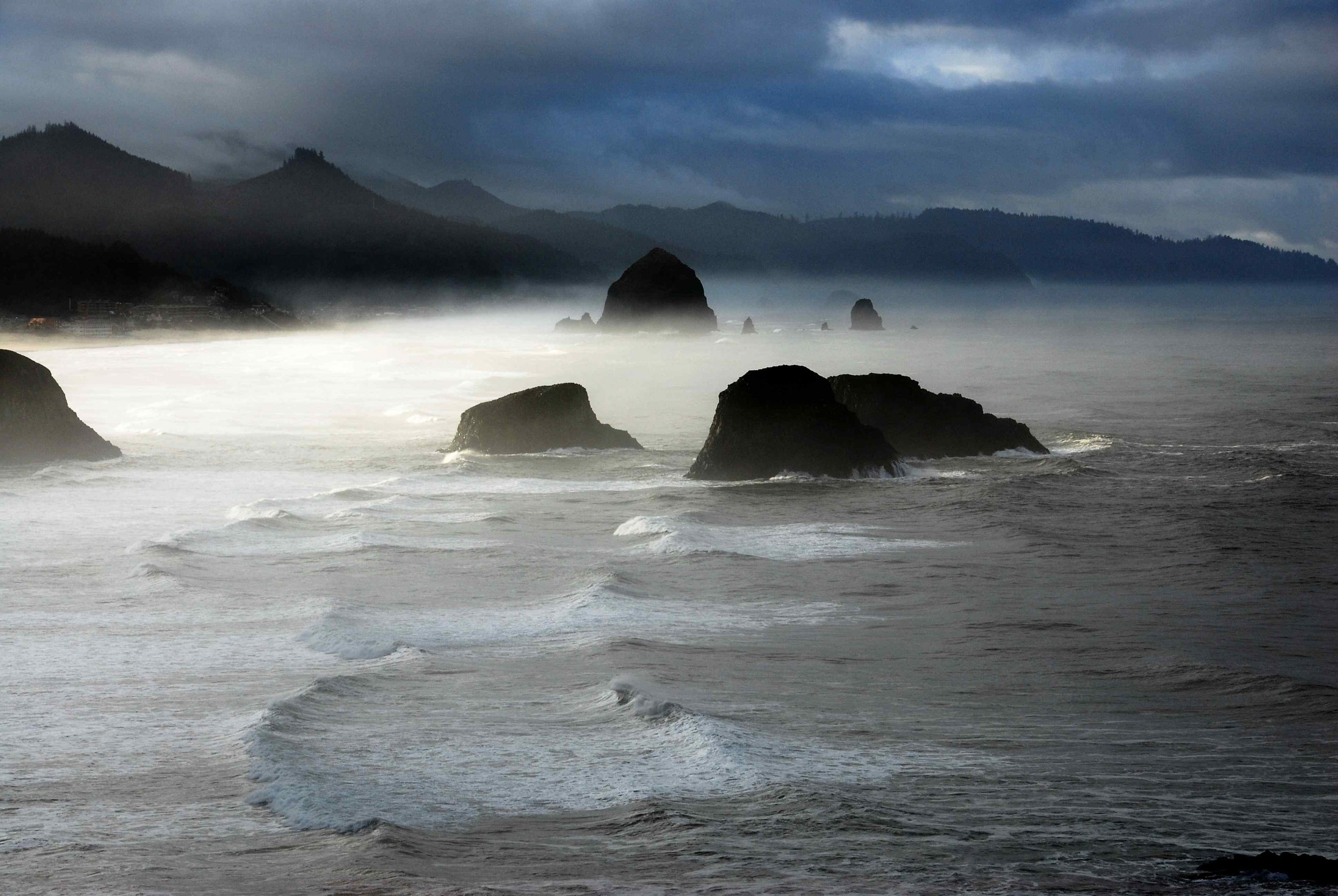 This photo captures a gloomy sunrise over Haystack Rock along Cannon Beach, Oregon. The photo is taken from Ecola State Park, with the iconic rock formation visible in the distance. The sky is cloudy and overcast, with shades of gray and blue dominat