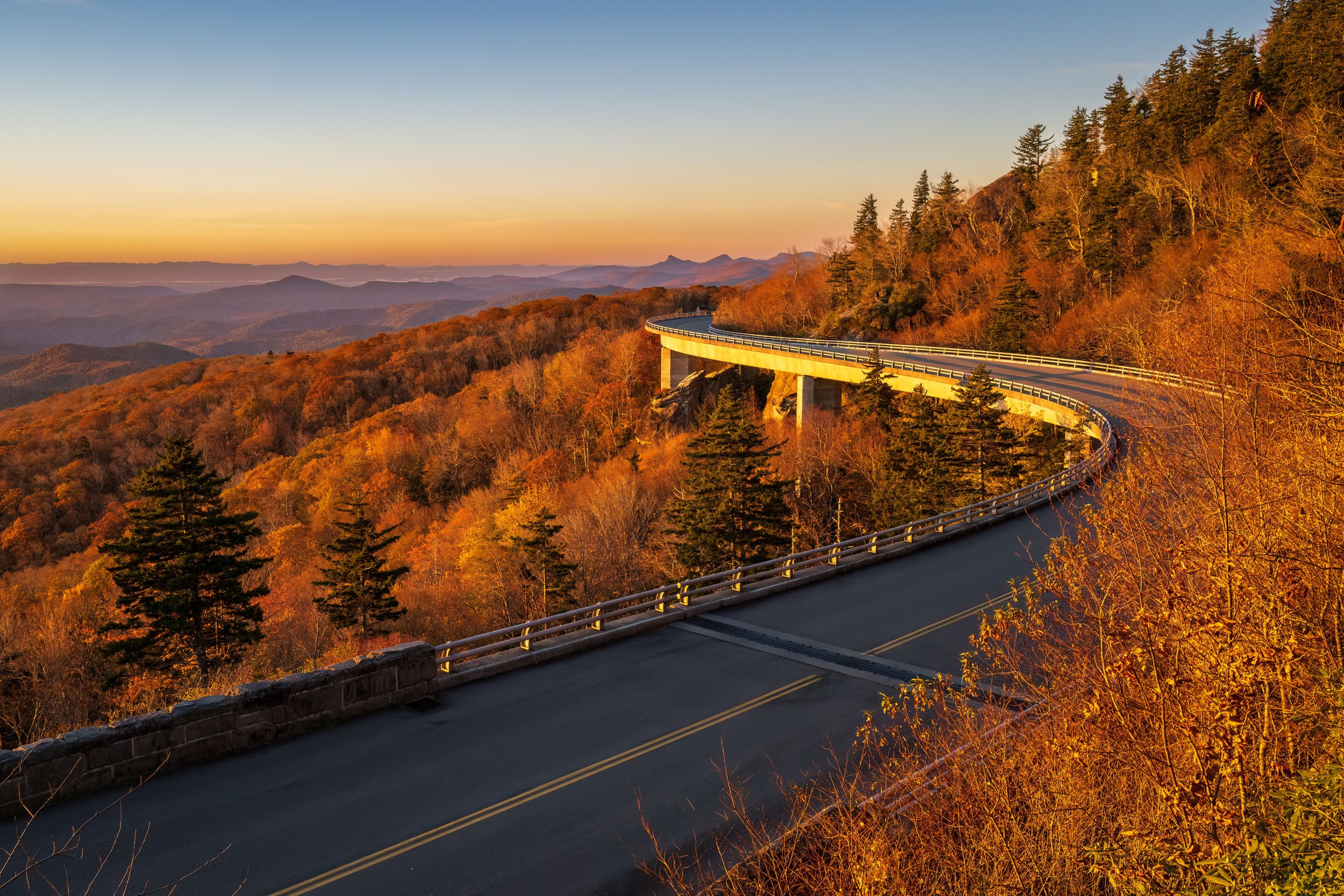 A warm fall sunrise across the Linn Cove Viaduct. The viaduct is seen stretching out into the distance, surrounded by a beautiful landscape of colorful trees and rolling hills. The colors of the trees range from vibrant oranges and yellows to deep re