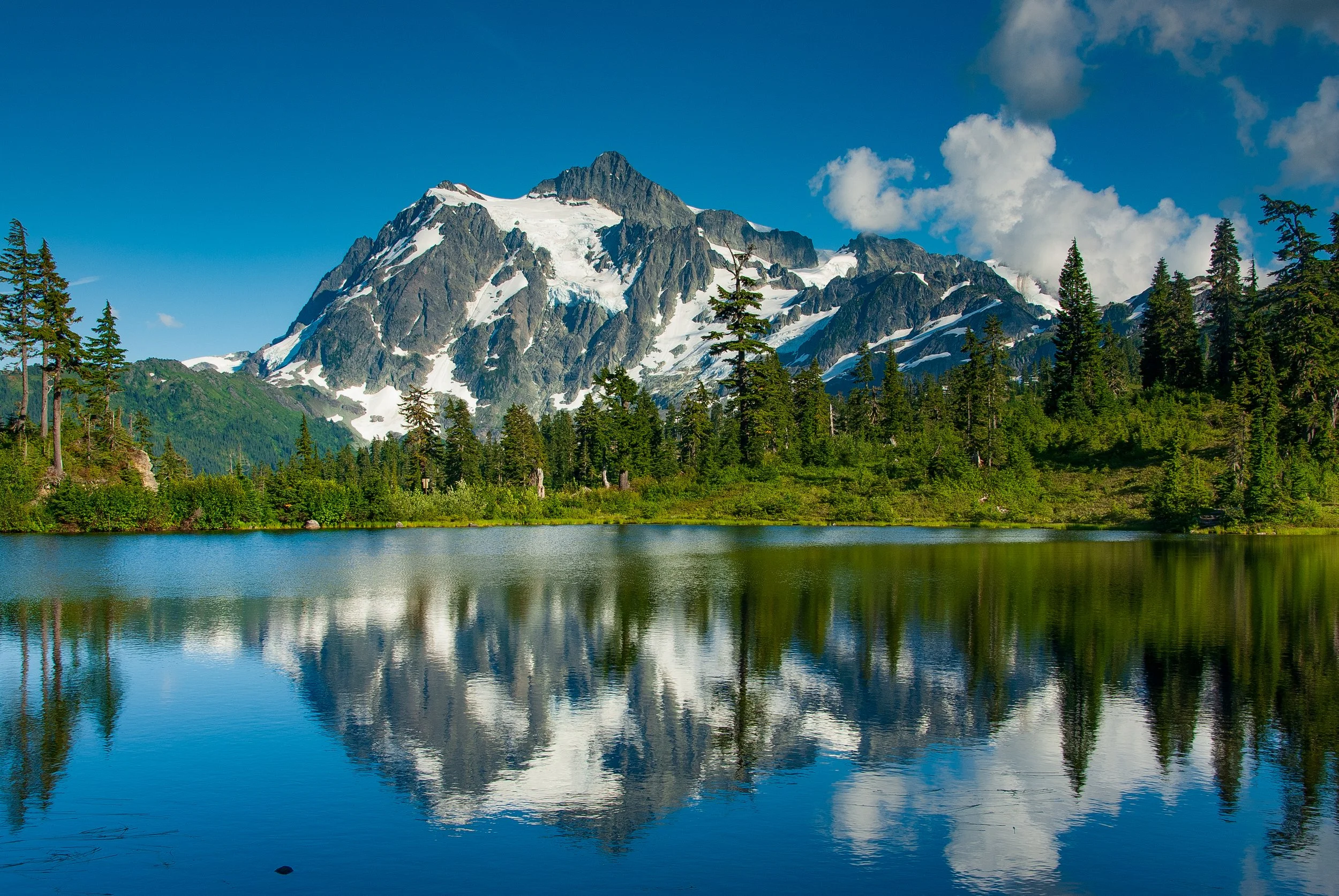 This photo captures the stunning beauty of Mount Shuksan and Reflection Lake. The mountain is viewed from a distance, with its distinctive peak standing tall against a clear blue sky. The reflection of the mountain in the calm waters of the lake crea