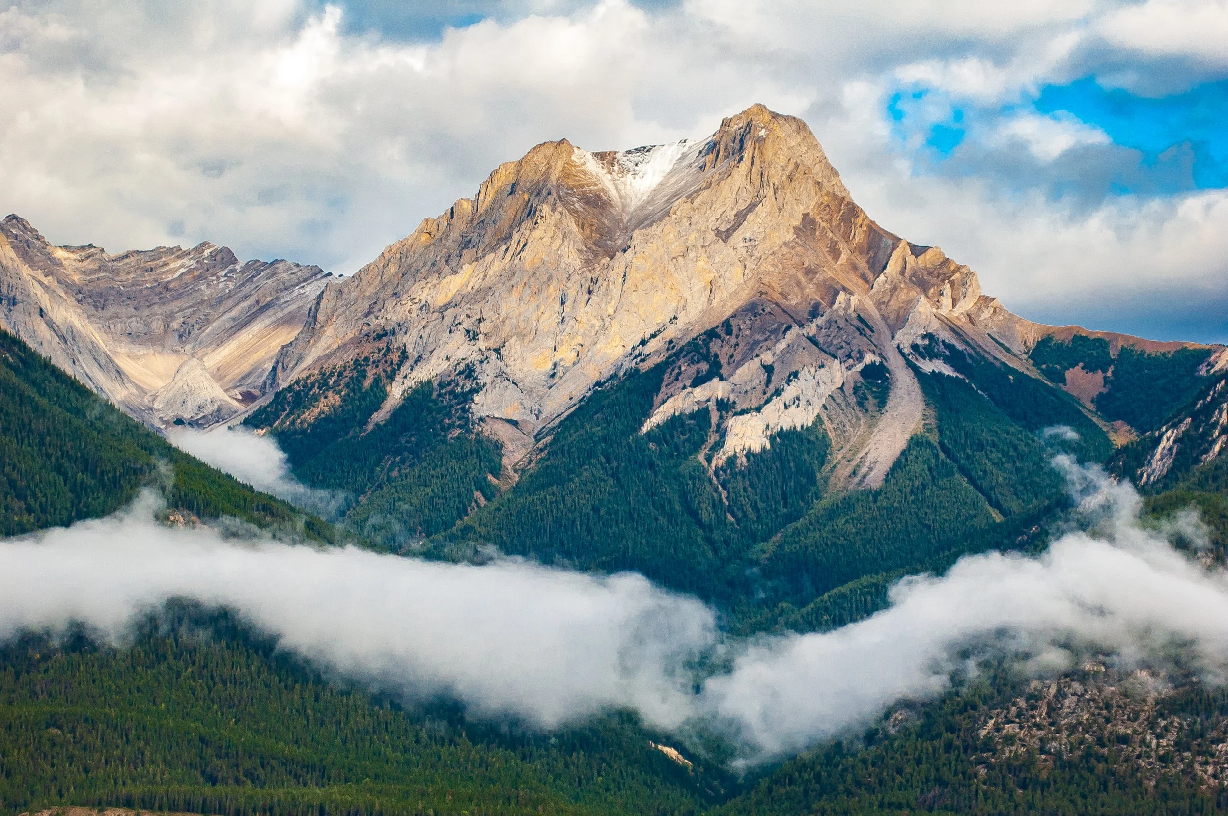 This photo captures the stunning beauty of center peak along the Three Sisters mountain peak in Alberta, Canada. The peak is viewed from a distance against a cloudy sky. The surrounding landscape is a mix of rolling hills and trees, with the mountain