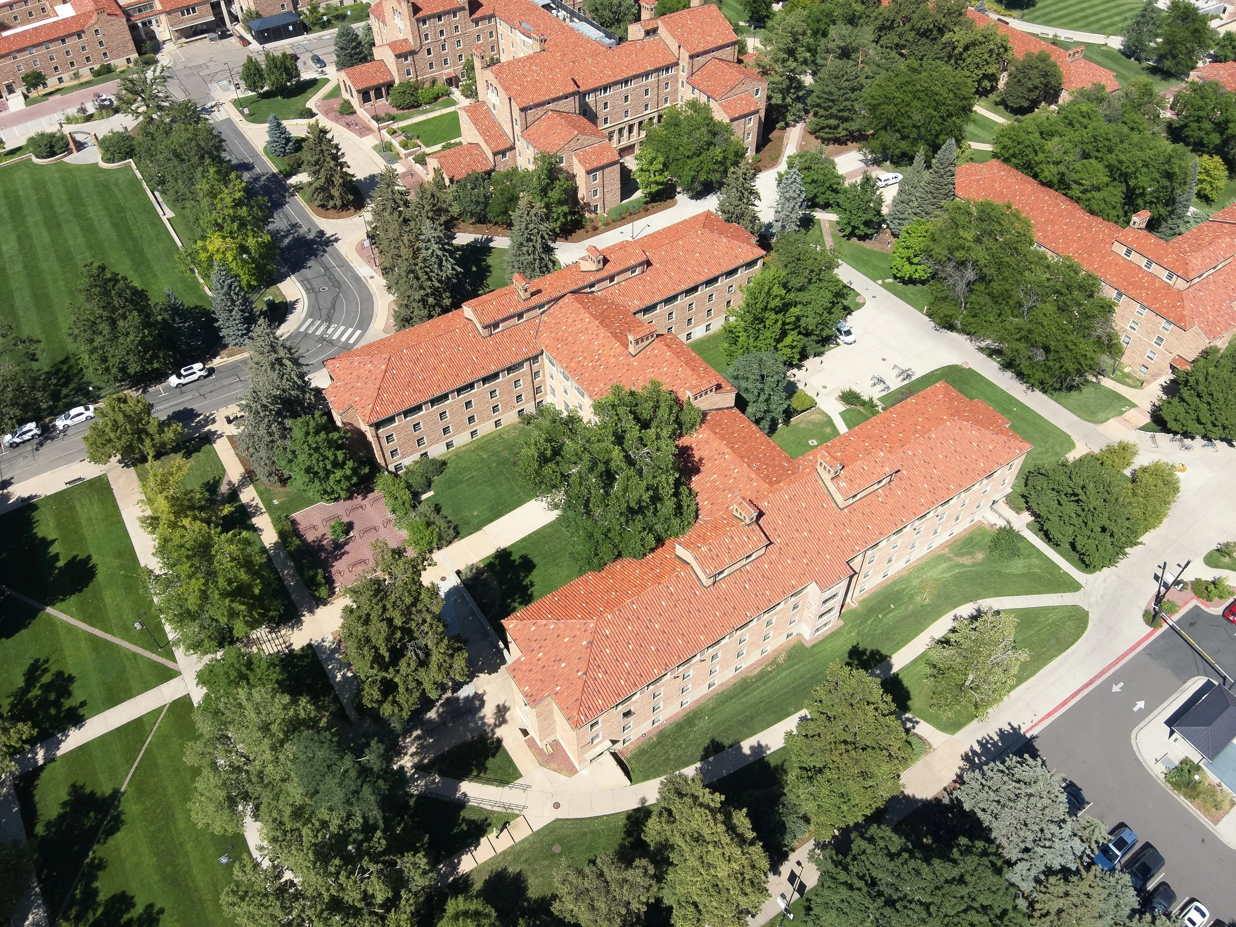 An aerial view of a residential neighborhood with multiple brick buildings, red-tile roofs, green lawns, and trees, with sidewalks and a street with cars.