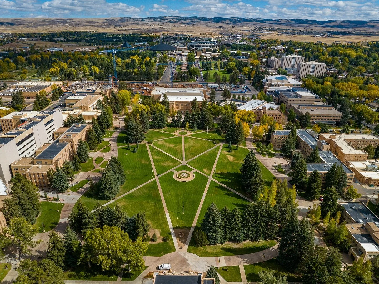 Aerial view of a university campus with green lawns, trees, pathways, surrounding buildings, and a cityscape in the background.