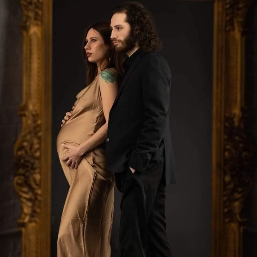 A pregnant woman in a satin beige dress standing next to a man in a black suit, both looking serious, standing in front of a decorative gold-framed mirror.