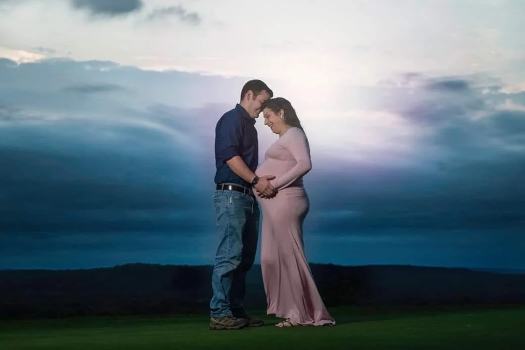 A couple stands close together for a maternity photo, holding hands, smiling, and touching foreheads against a landscape background with a cloudy sky.
