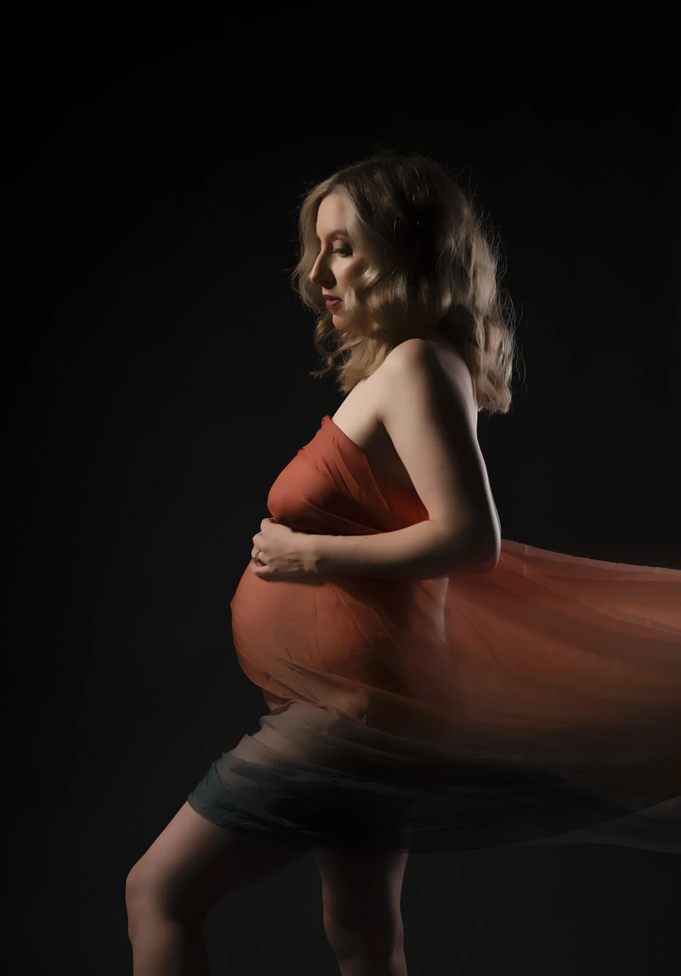A pregnant woman with wavy brown hair posing against a black background, wrapped in a flowing, semi-transparent orange fabric.