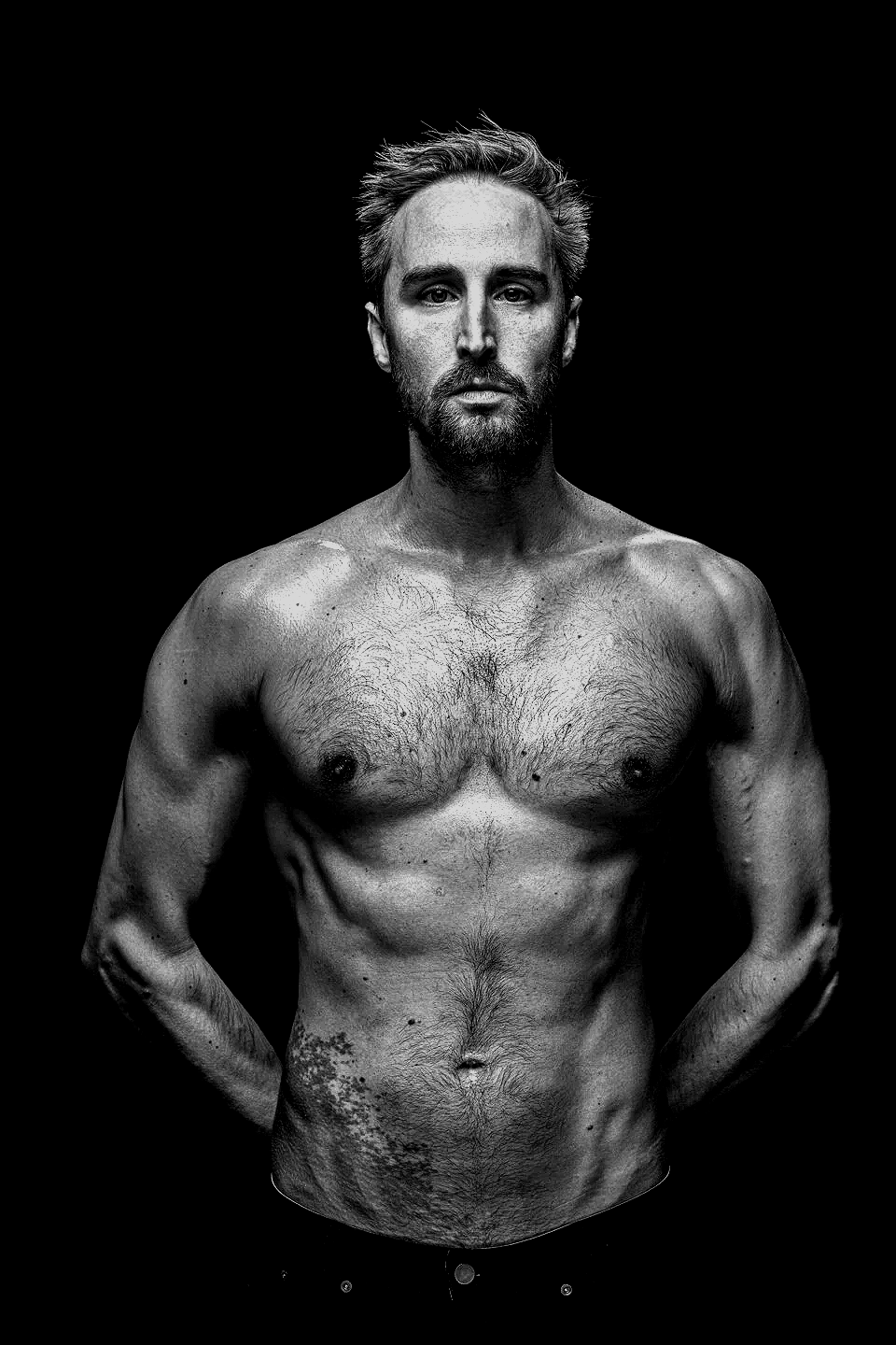 Black and white portrait of a shirtless man with short hair and facial hair, standing against a dark background.