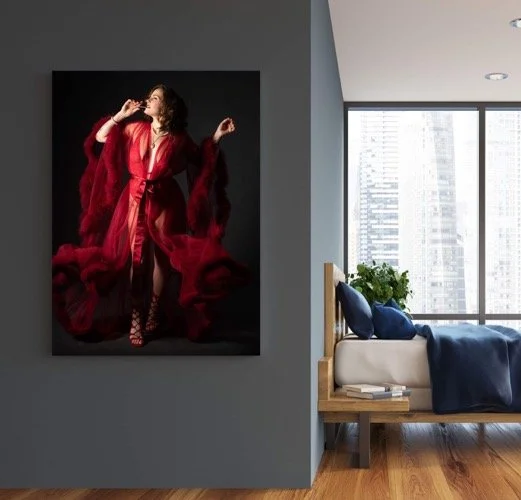 A modern bedroom with large windows showing city skyscrapers, a bed with blue bedding, a plant, and a large artwork of a woman in a red dress on the wall.