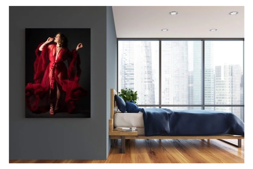 Modern bedroom with large windows overlooking city skyline, wooden floor, bed with navy blue bedding and pillows, plant on bedside table, large artwork of woman in red dress hanging on wall.