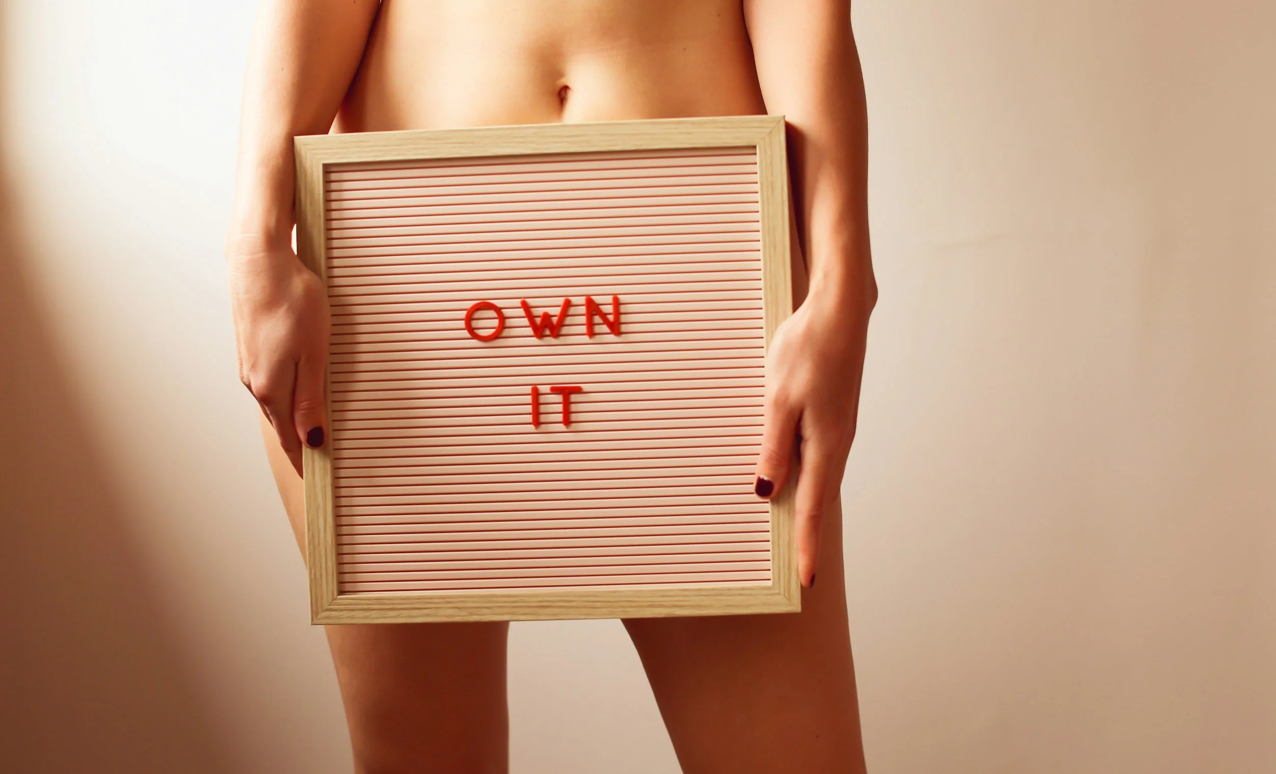 A person holding a pink felt letter board with the words 'OWN IT' spelled out in red letters, standing against an off-white background.