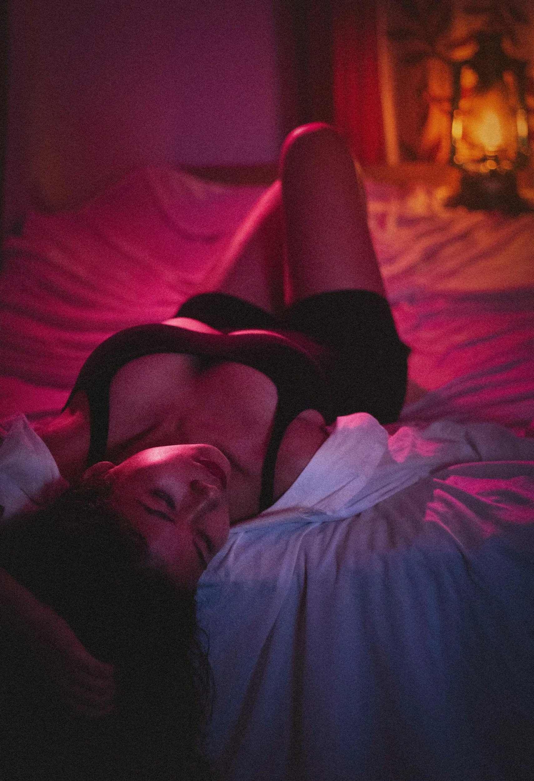 A woman lying on a bed with pink and purple lighting, smiling and wearing a white shirt and black underwear, with a woman in the background sitting near a lamp.
