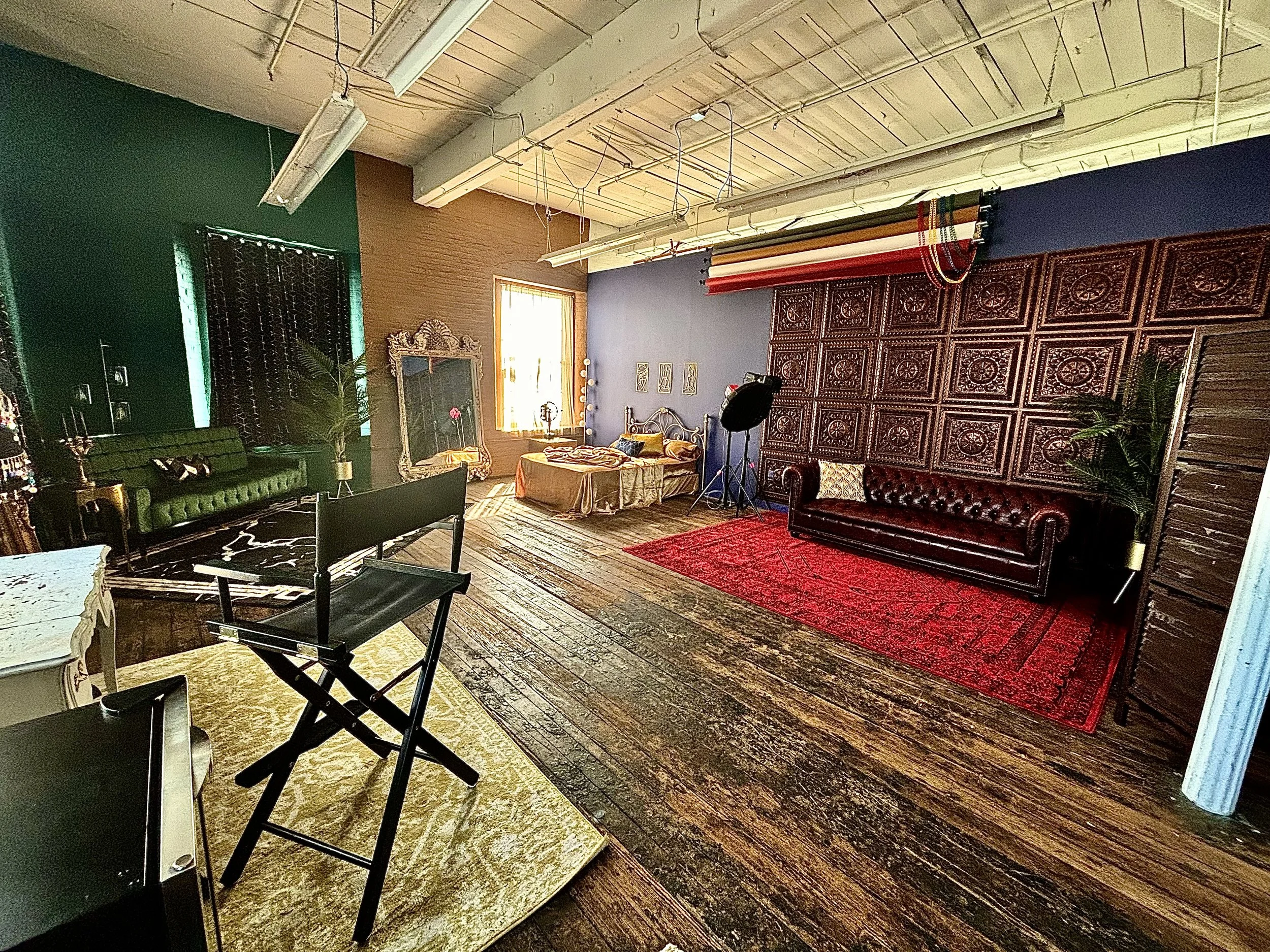 Interior of a room with eclectic decor, wooden floors, a green sofa, a vintage mirror, a purple accent wall, and a large ornate wooden wall panel with decorative elements. There are various tables, chairs, and a studio light setup.