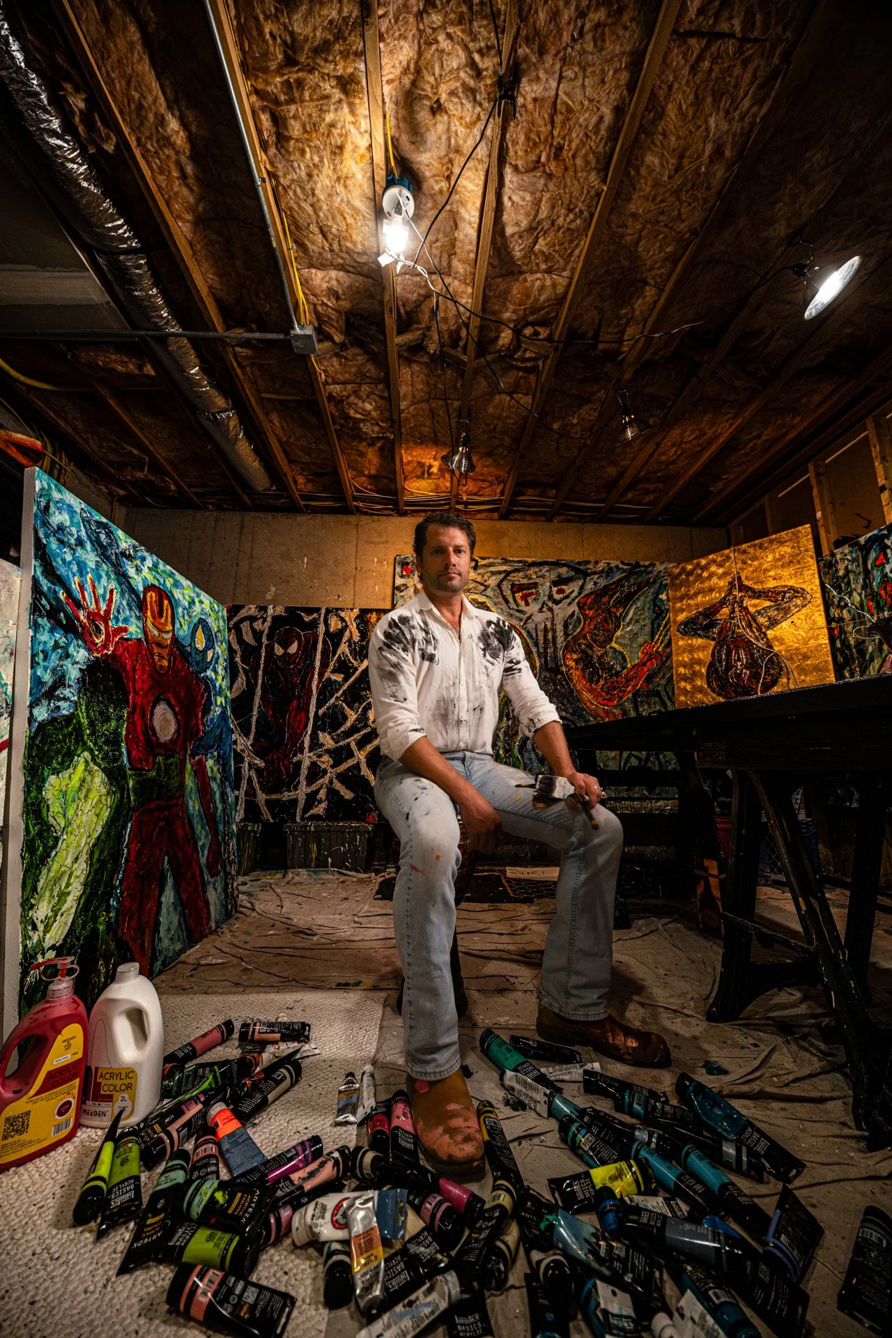 An artist, wearing a white shirt and paint-stained pants, sits on a chair in an art studio with colorful paintings behind him. The studio has a wood-paneled ceiling with exposed insulation and is scattered with paint tubes, bottles, and art supplies 
