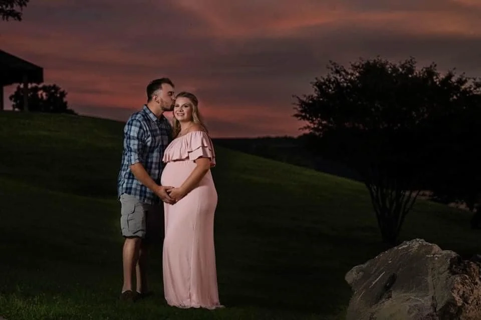 A maternity photo of A pregnant woman in a pink dress and a man in a blue plaid shirt and shorts standing close together outdoors at sunset, holding hands and smiling.